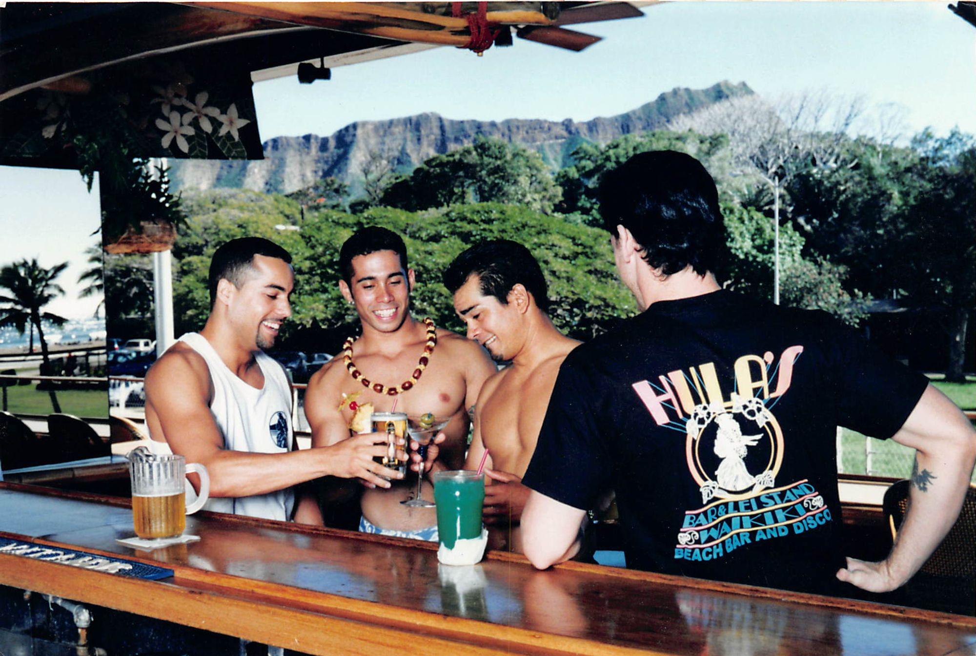 Three men cheers in a Hawaii bar, amidst a mountainous tropical landscape. Another man faces them, his back turned towards the camera, wearing a shirt that says "HULA'S BAR & LEI STAND"."