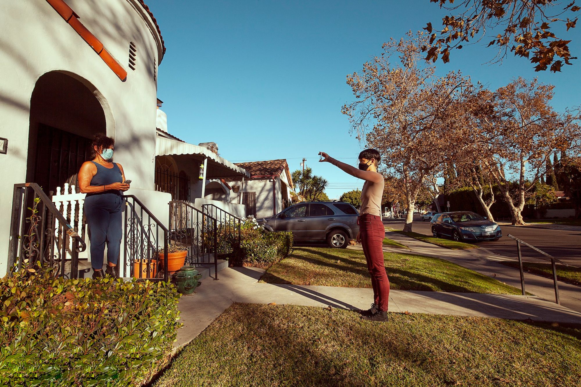 The artist wears red pants and a beige shirt, and stands up straight while holding up her smartphone with an outstretched arm. She faces a residential house, where a woman in a blue tank top stands in the entryway to what appears to be her home. The two women face each other, with a blue sky in the background.