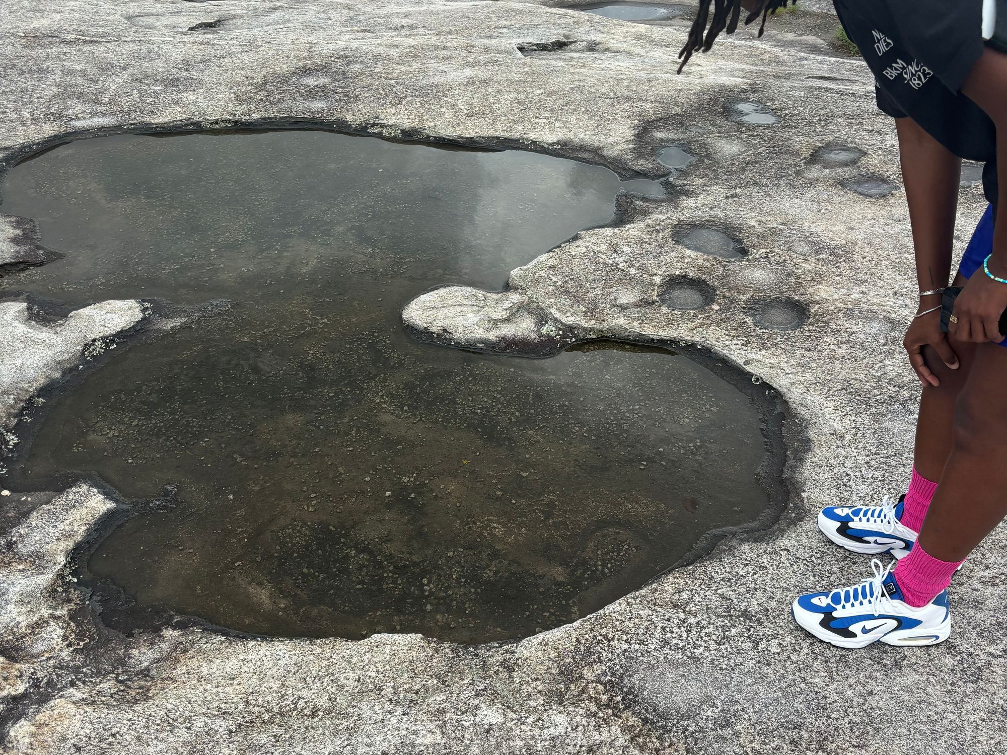 An iPhone photograph of a figure kneeling towards the small pools that form between rock ridges on Stone Mountain