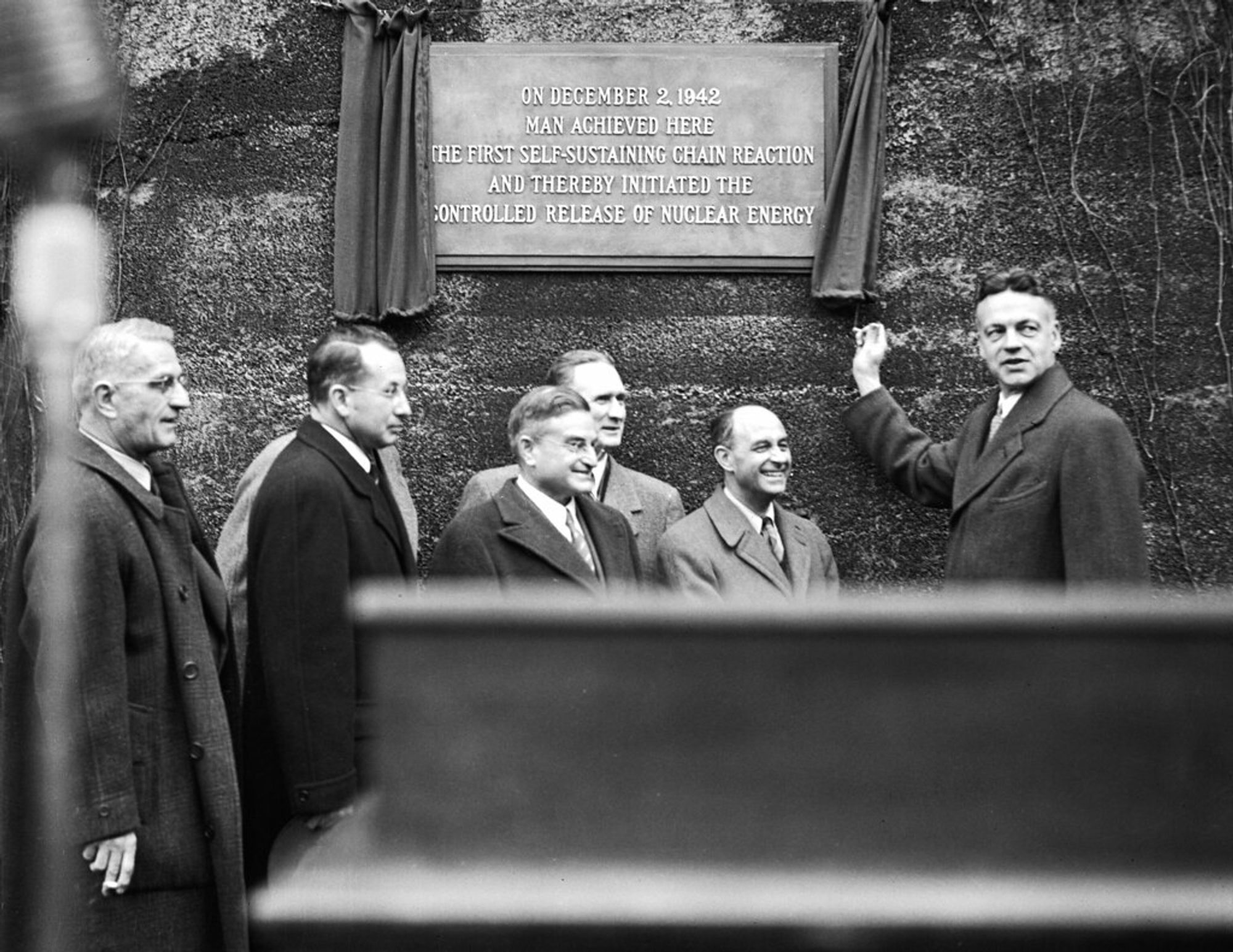 A balck and white photo of scientists in front of a plaque honoring the nuclear chain reaction december 2nd, 1942.