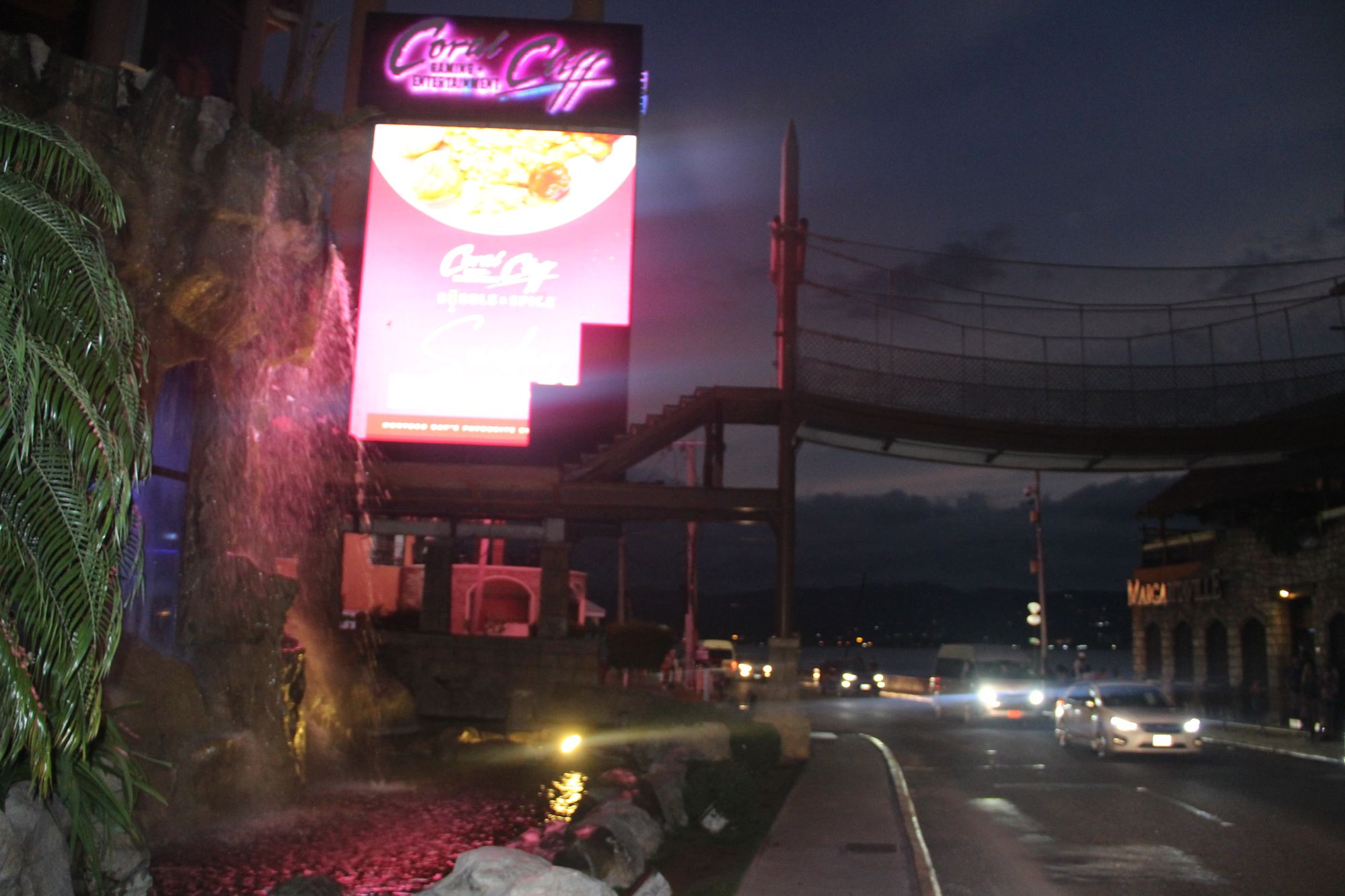 A relatively blurred image of Montego Bay at night, with cars passing by a strip, replete with a Gaming and Entertainment center called "Coral Cliff," lit up in pink.