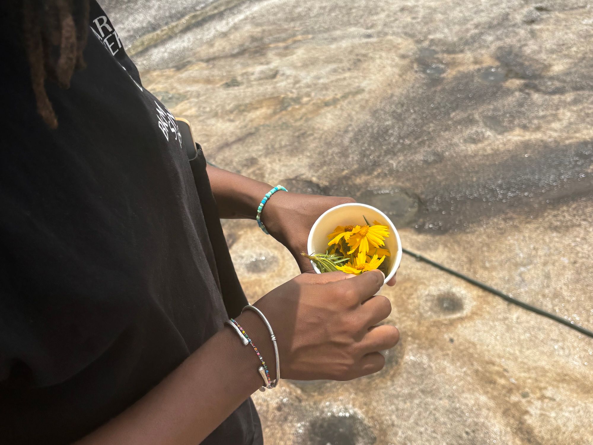 An iPhone photograph of a figure, only seen in profile, who holds a paper cup filled with yellow flowers and stands on what looks like a granite ground.