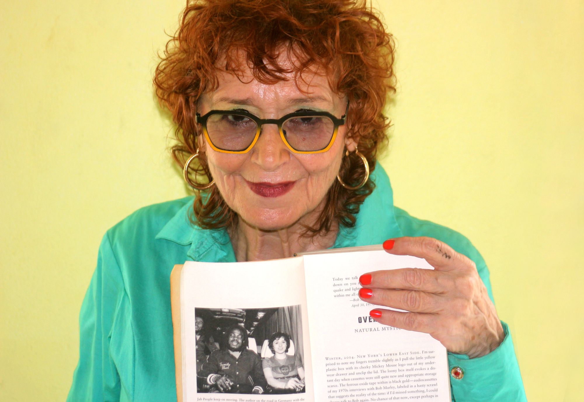 An older woman wearing black and yellow framed glasses, holding a book open to a page with a black-and-white photograph of two people sitting side by side. 