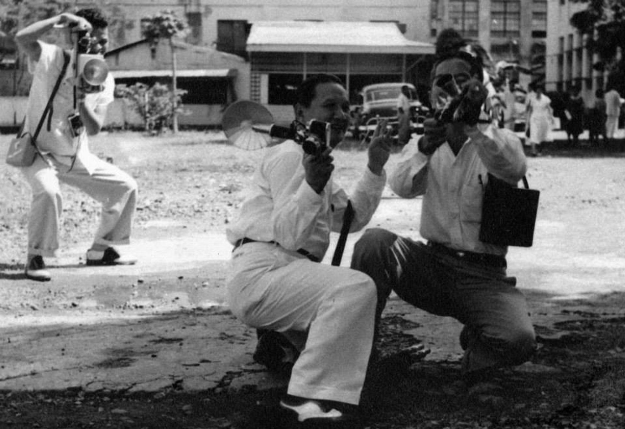 A black-and-white photograph of Protomartir and another photographer crouched on the street and holding their cameras. They are wearing button-up shirts with slacks and are looking in the same direction.