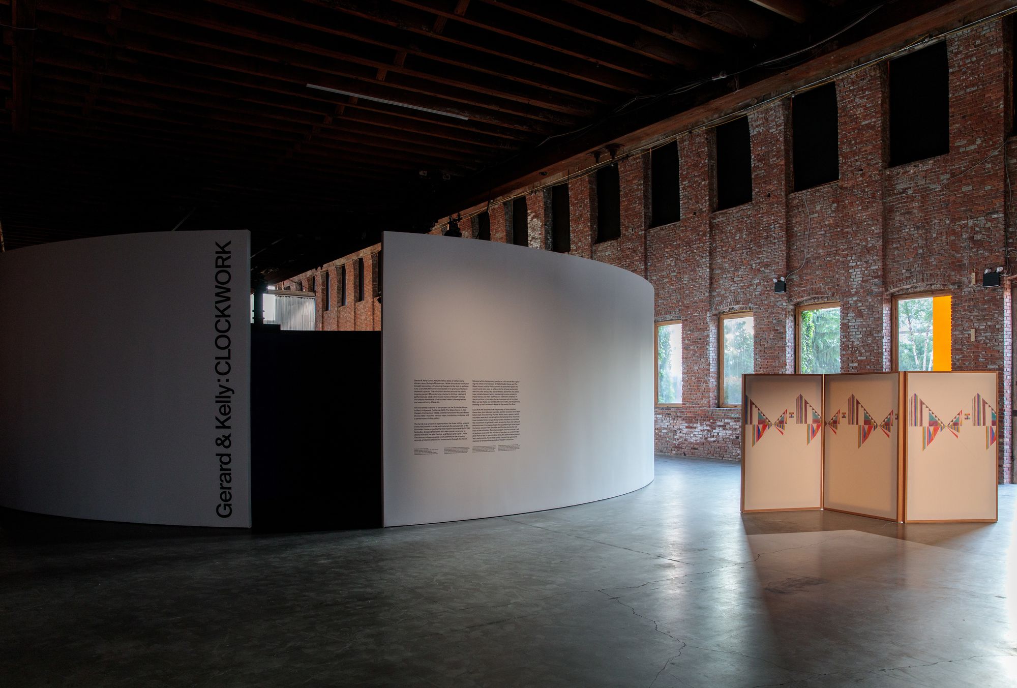 An installation view of the entrance of CLOCKWORK, featuring the entrance to the round-walled video room to the left, and a human-sized folding screen painted in pastels to the right.