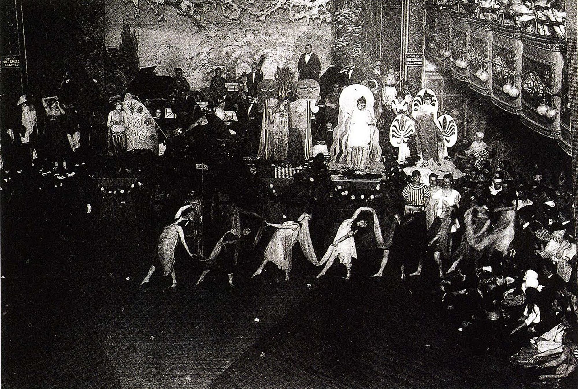 A black-and-white image of costumed figures dancing around a stage for a packed audience.