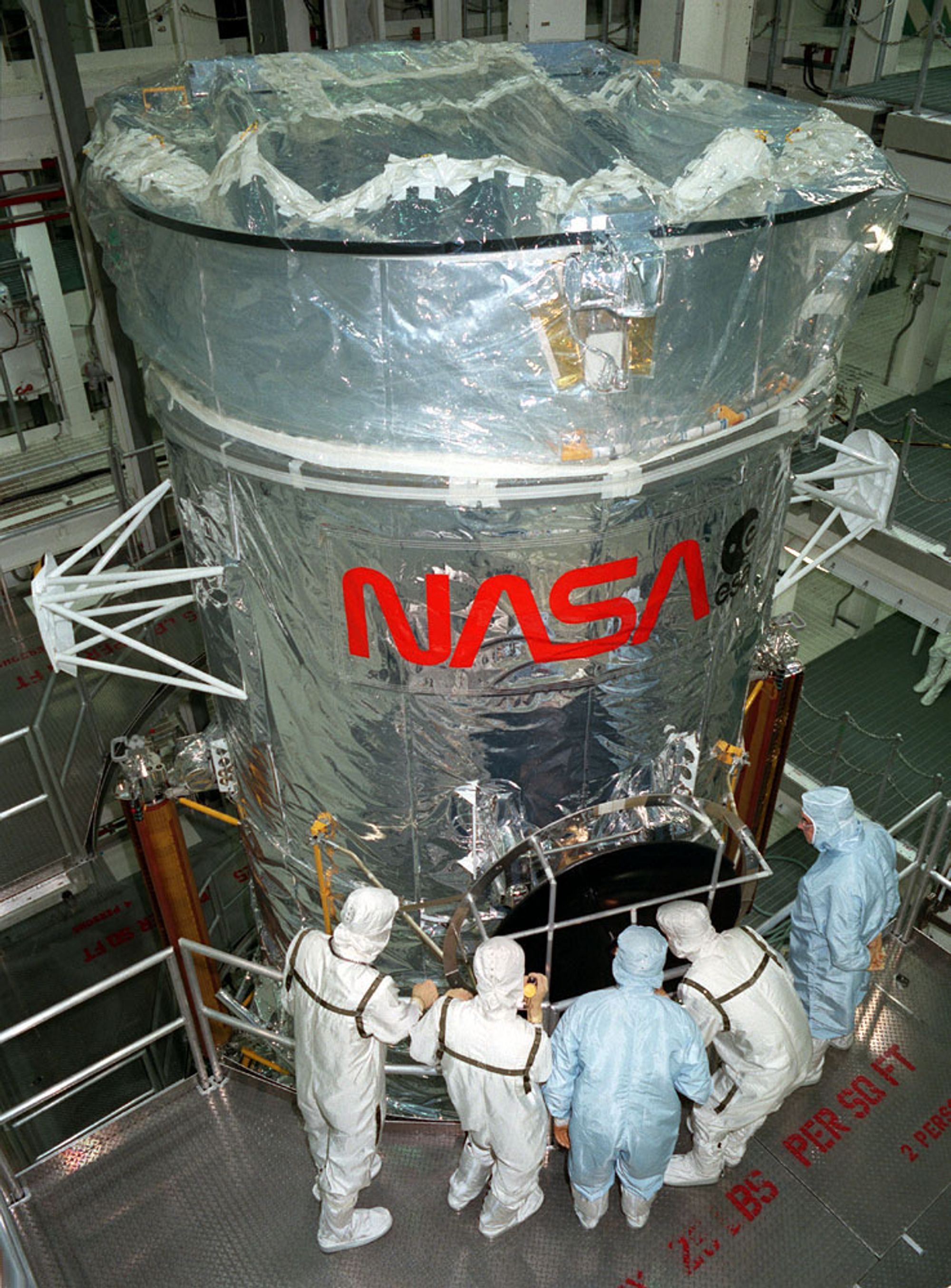 Five men in white and blue suits gaze into the darkened base of a telescope, covered in a silver material with a red NASA logo wrapping around its front.