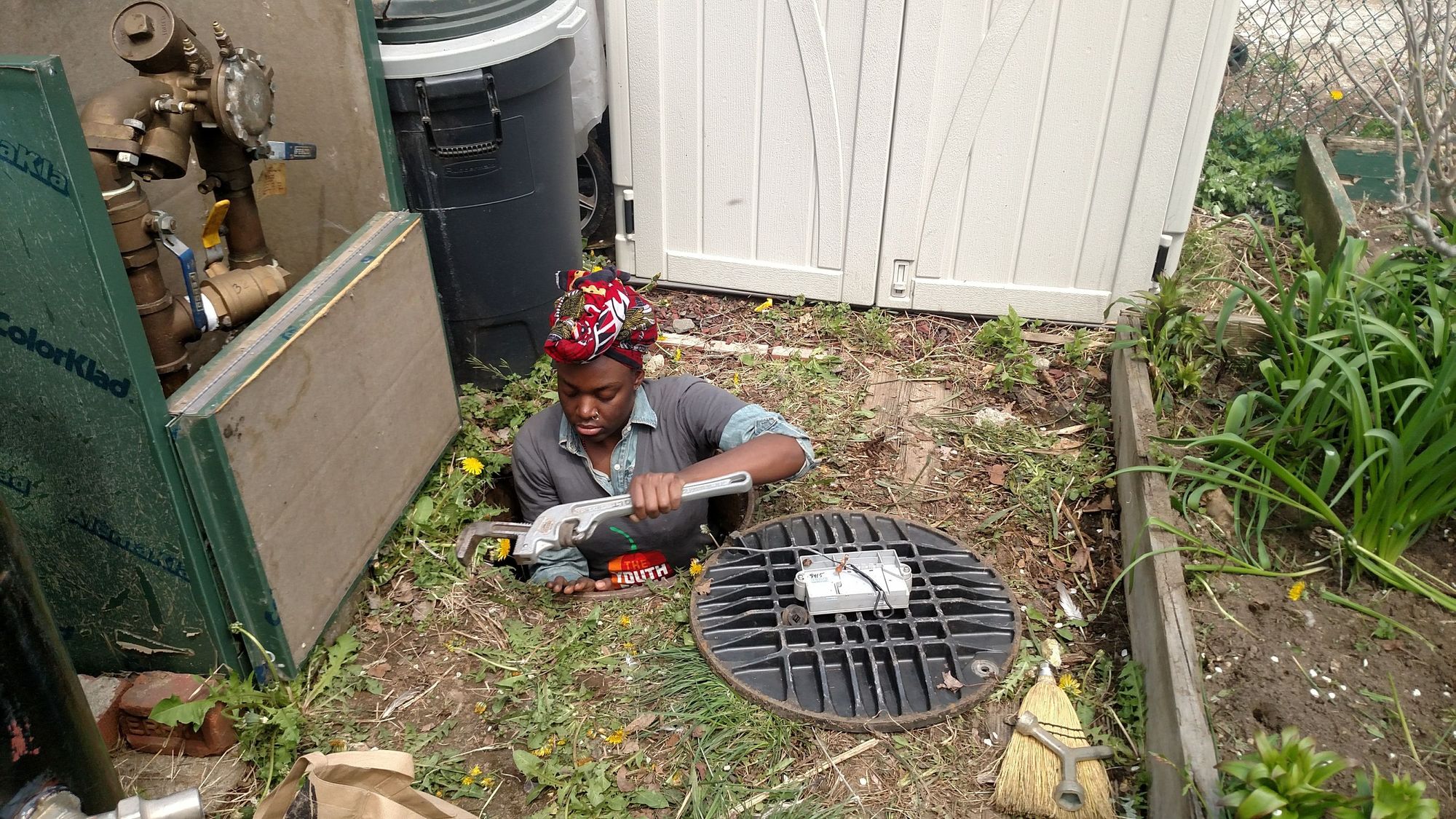 Sawdayah Brownlee performing water systems maintenance in a manhole in a garden, from the Merrick-Marsden Neighbors Association Garden, a BQLT garden, Jamaica, Queens