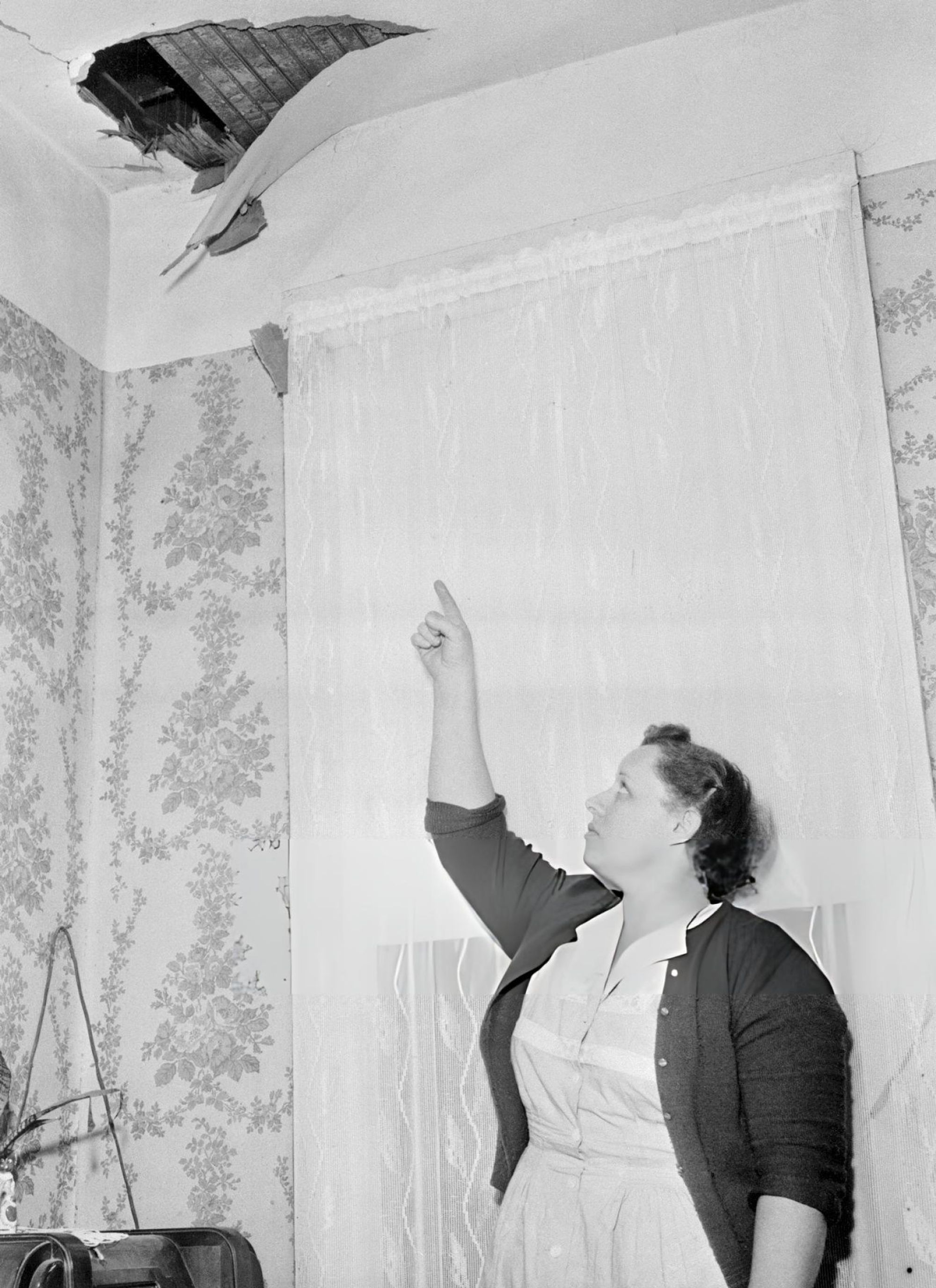 A woman dressed in a 1950s house dress points to a hole ripped through her ceiling. 