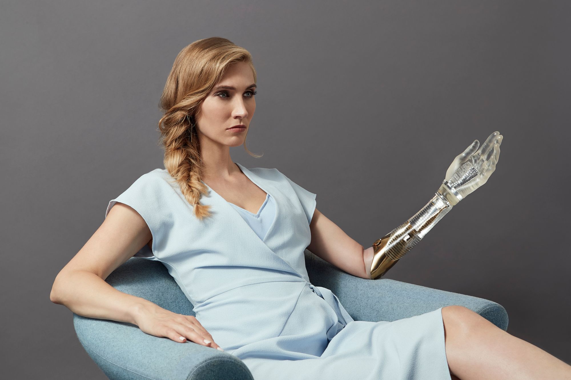 A blonde white woman is showen in a light blue dress. She extends her arm out and is modeling a prosthetic arm and hand that is glassy and semi-opaque.
