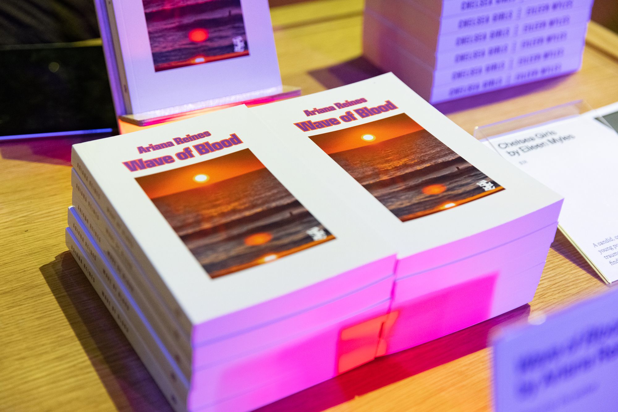 A stack of copies of "Wave of Blood" by Ariana Reines. The book cover features the author and title at the top, with an image of a sunset over waves. 