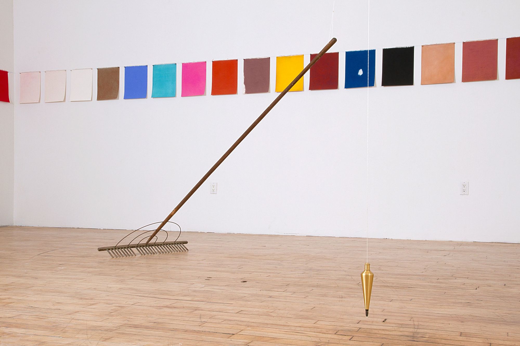 An installation view of Kathleen White's "A Rake's Progress," which features a long row of colorful, paper sheets, and a hanging plumb and rake in the middle of the floor
