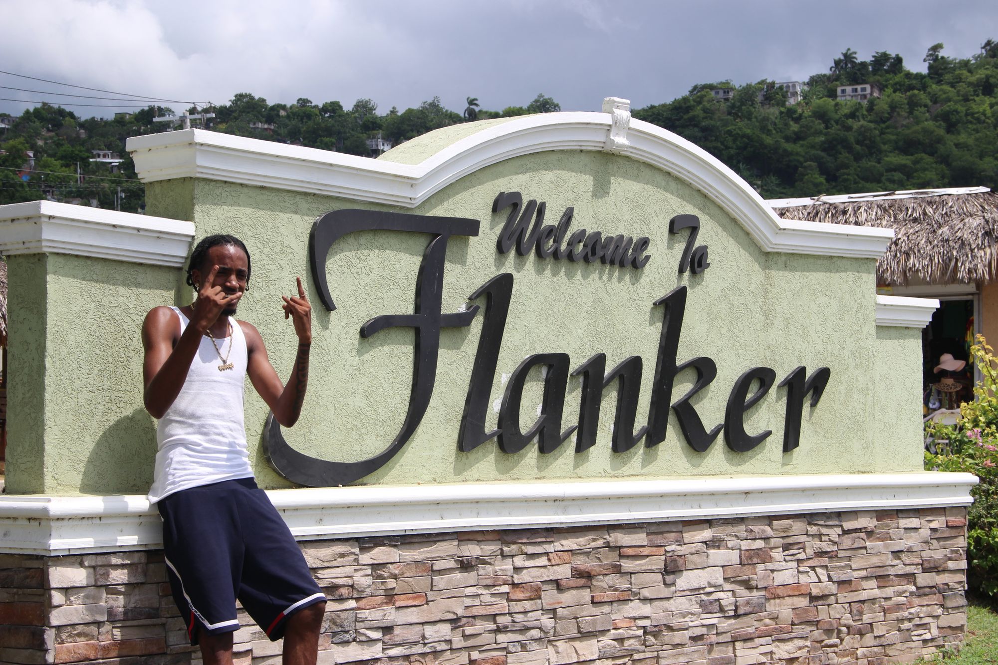 A man leans against a large sign that reads "Welcome to Flanker" and squints his eyes in the sun, holding up hand signs.