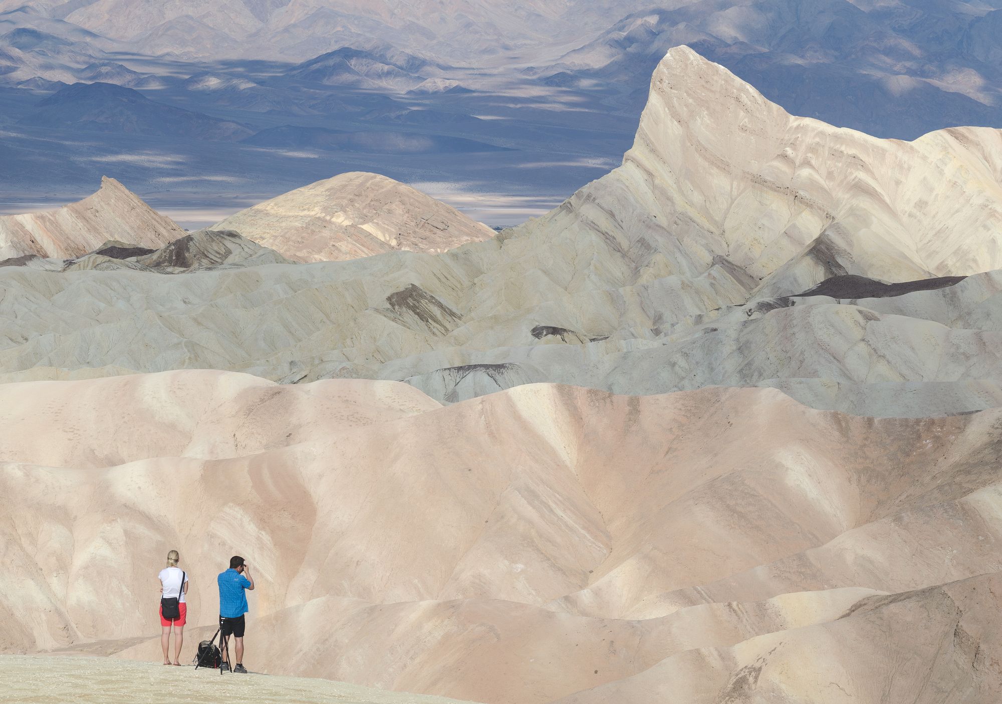 Two small figures stand on a ledge and photograph the beige peaks of Death Valley. 