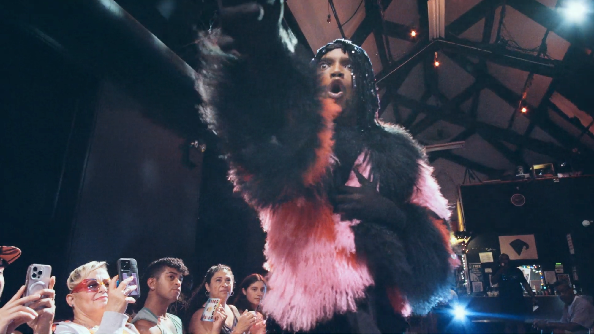 A person in a pink and black fur coat with black gloves and hands stretched out in front of a crowd at a ball show