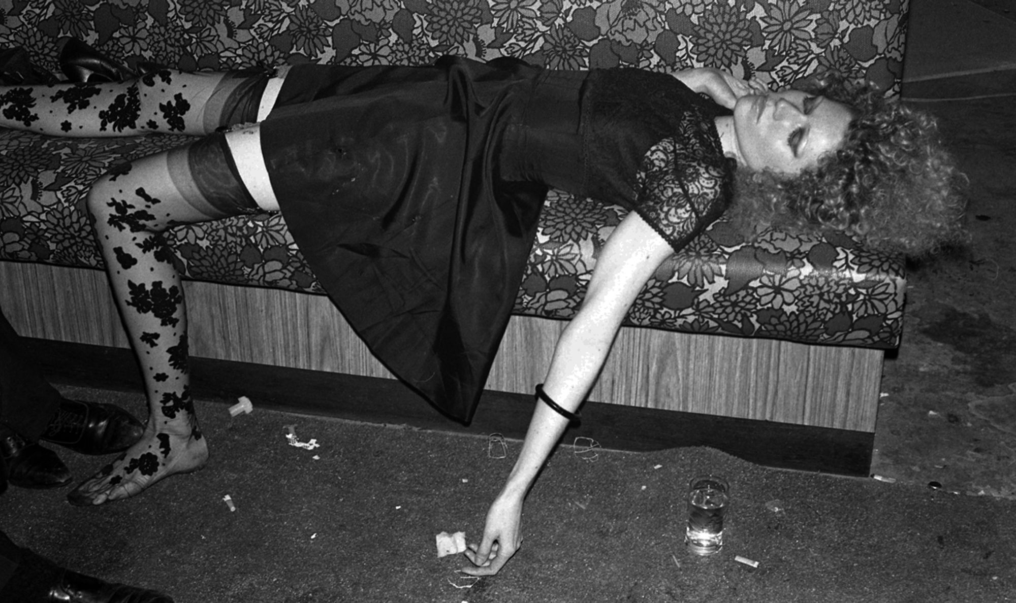 A woman wearing a black satin dress and black lace stockings lies unconscious on a couch.