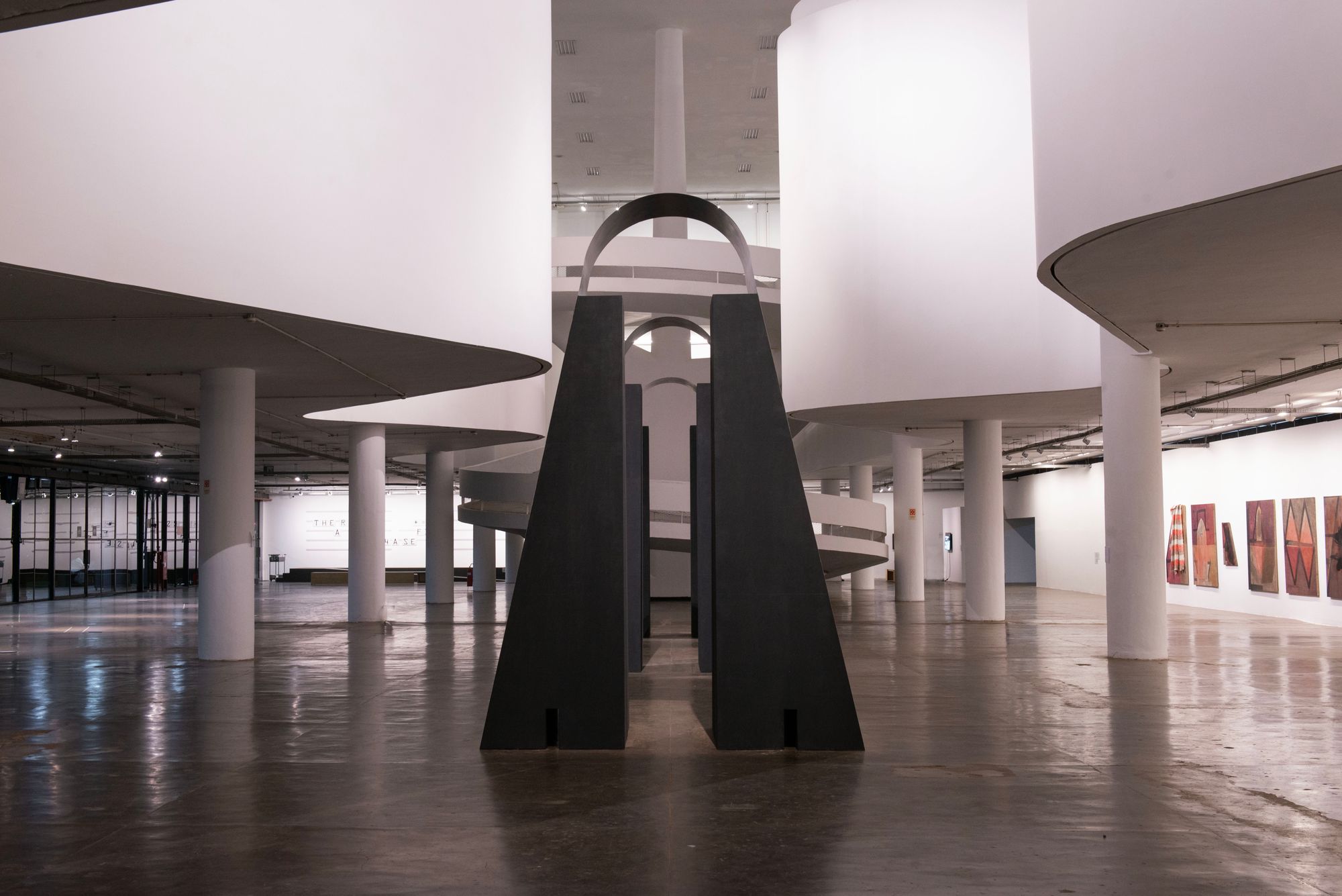 A winding gallery space with art on each wall and a meandering staircase intersecting the center of the image, where a large metal sculpture rests and bisects the space.