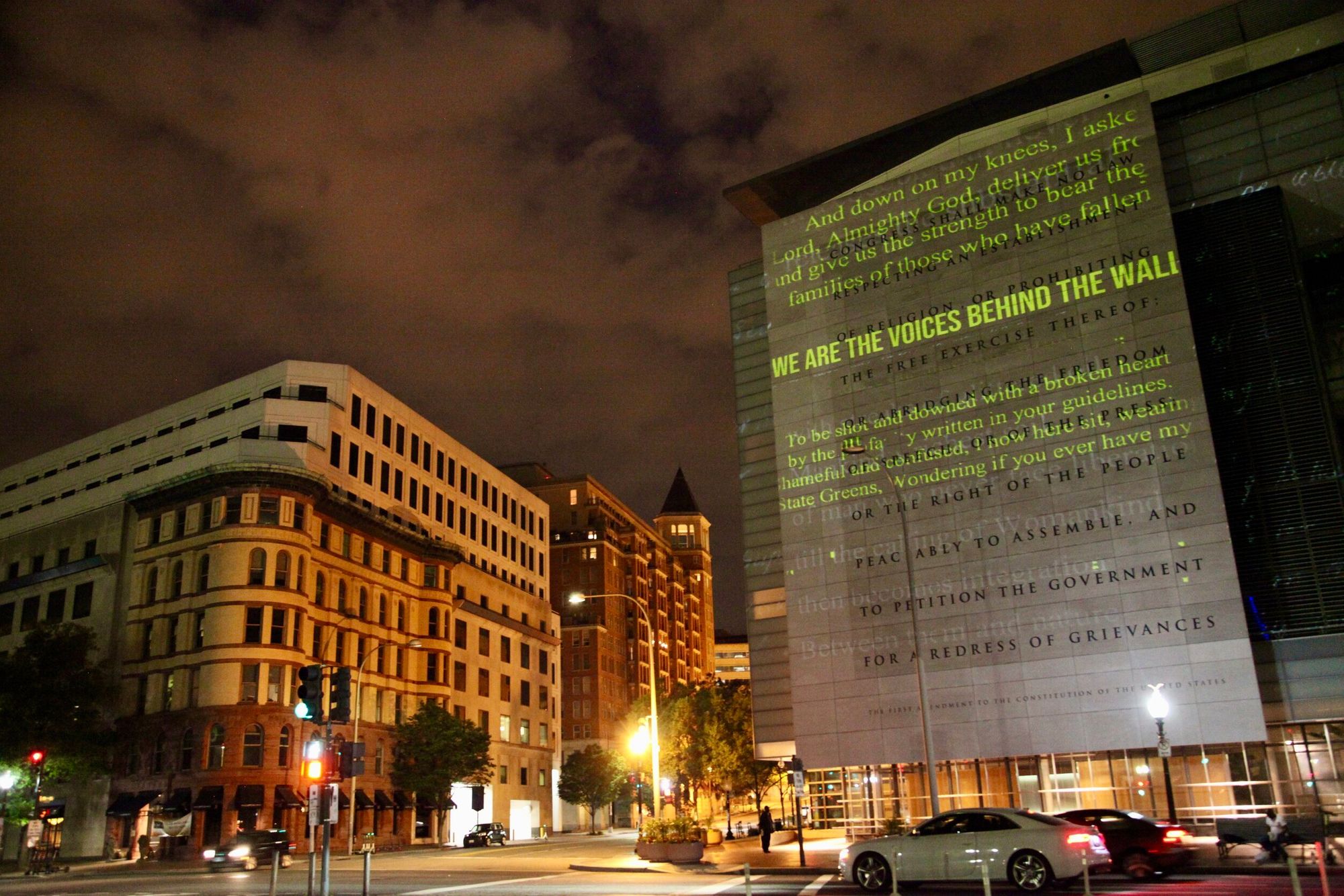 A street scene set at night that shows two buildings. On one building to the right, there is a large projection of text that scales the entirety of the building. The text reads "WE ARE THE VOICES BEHIND THE WALL."