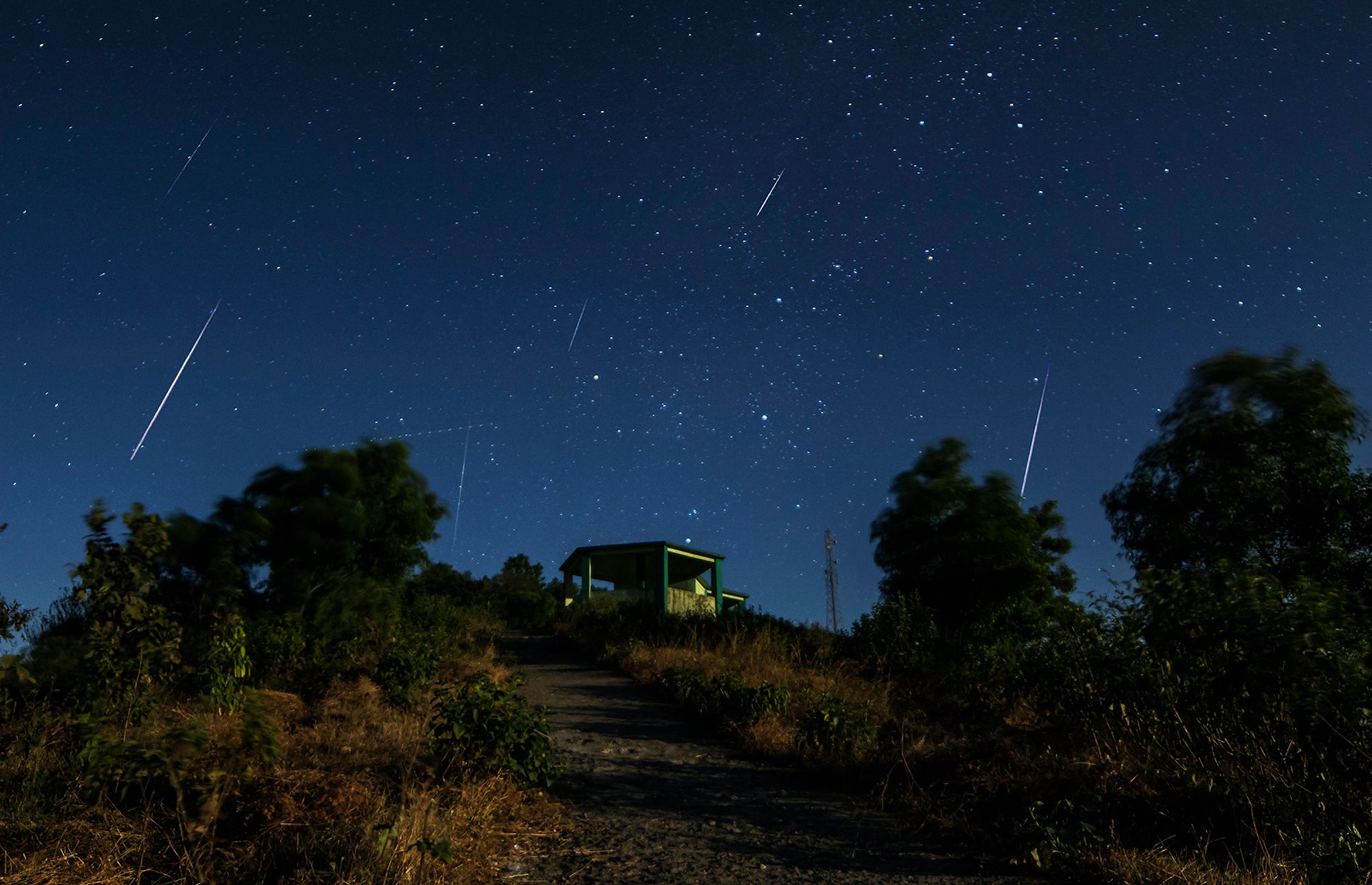 A structure on a hill is surrounded by a nigh sky full of streaking meteors.