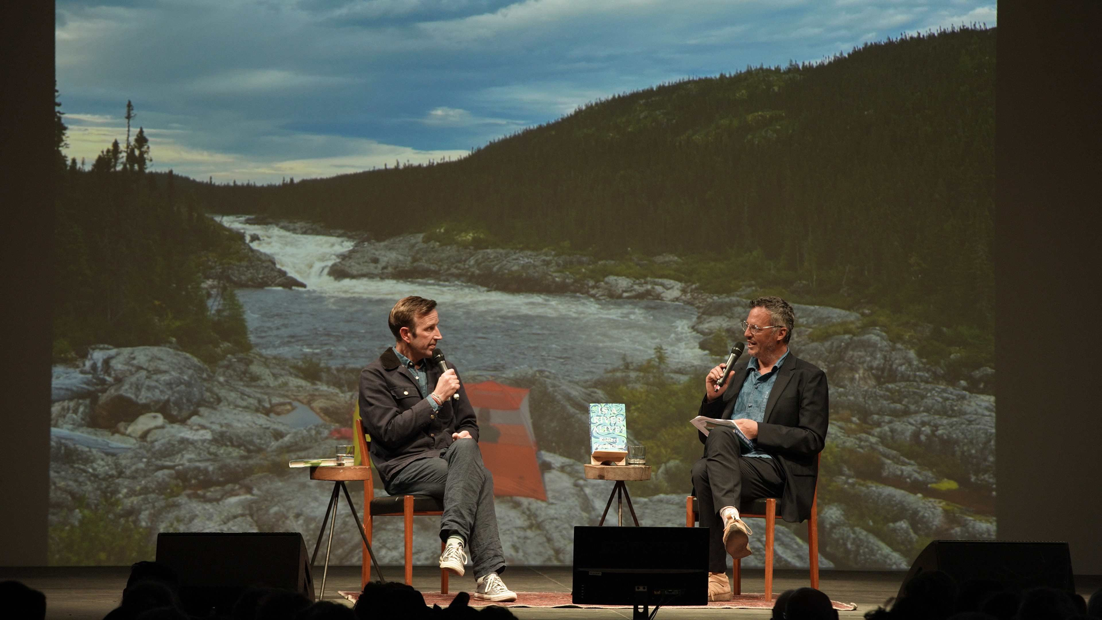 Two men sit on a stage against an image of a river, speaking into microphones.
