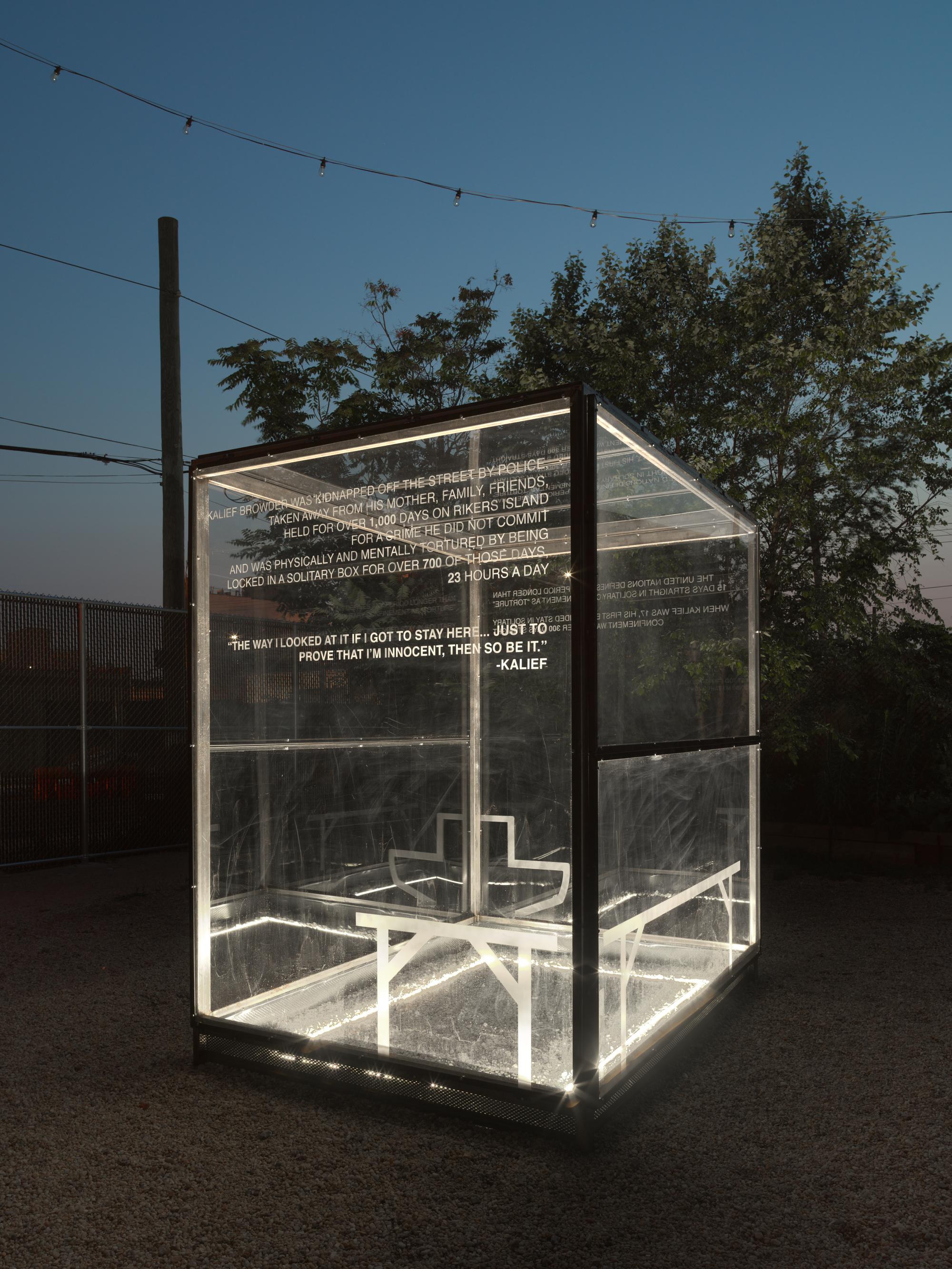 A glass sculpture of a box that replicates that parameters of a solitary confinement cell. The sculpture is lit from below and it is dark outside. 