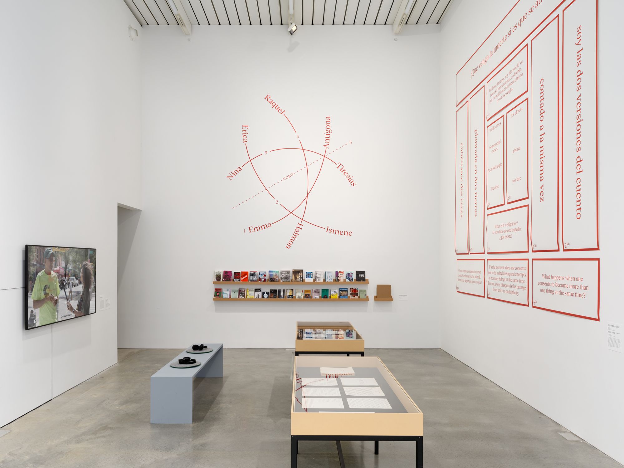 A gallery space with a couple of tables, and charts in orange ink marking the walls in Spanish and English