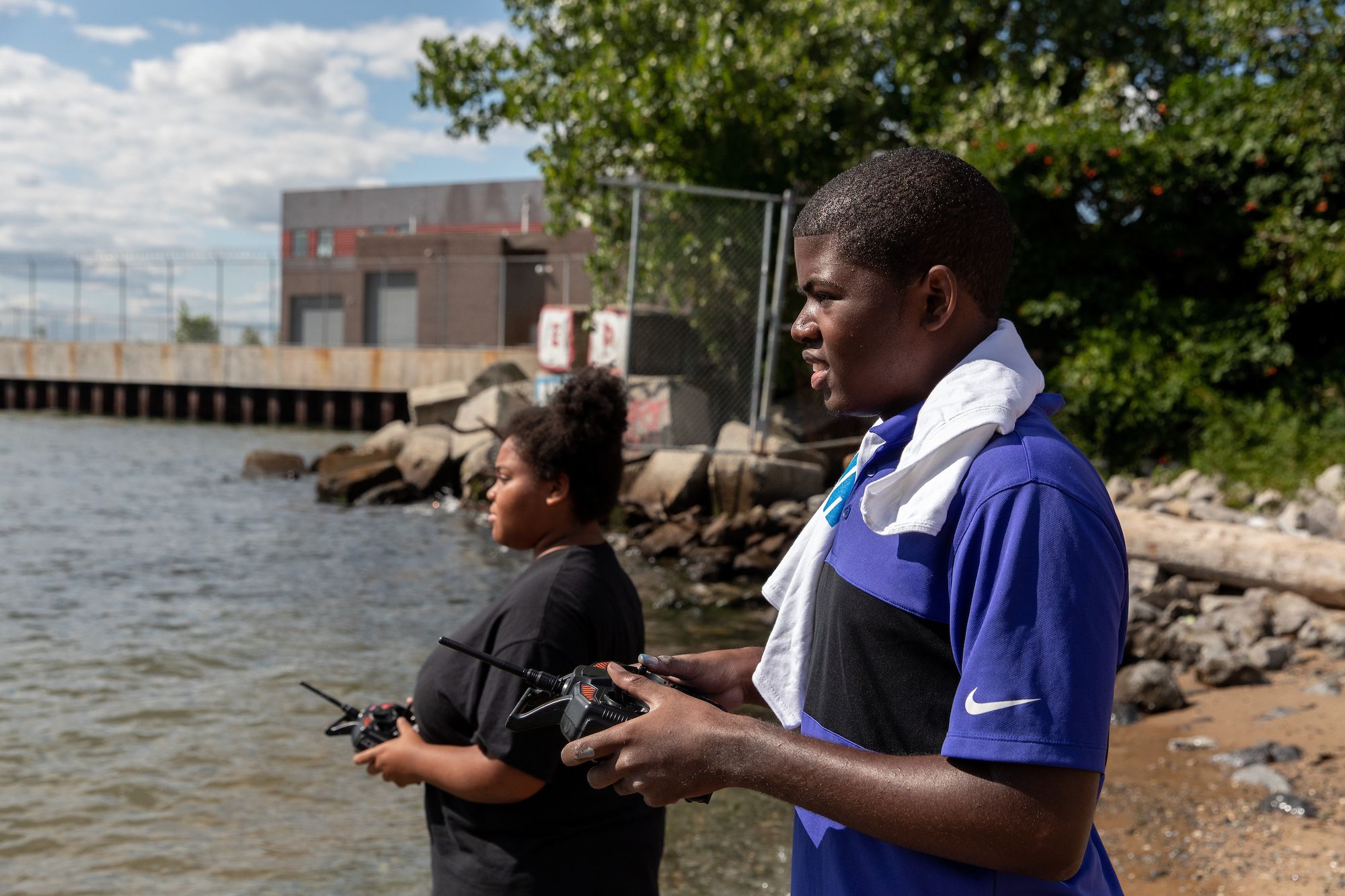 Two teens stand on a modest beach and look out over the water as they drive their boats via remote control controllers.