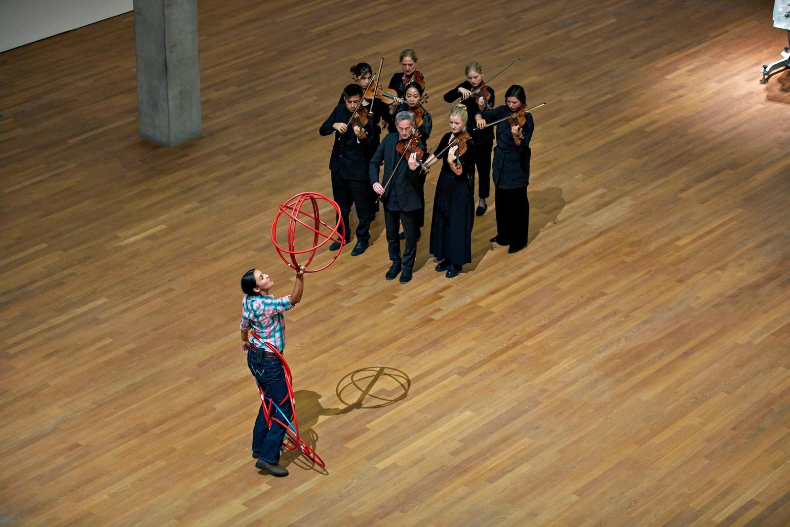 On a wooden performance floor, Bigstone Cree Nation dancer Sandra Lamouche spins a red hula hoop ensemble while a small orchestra group dressed in black plays the violin