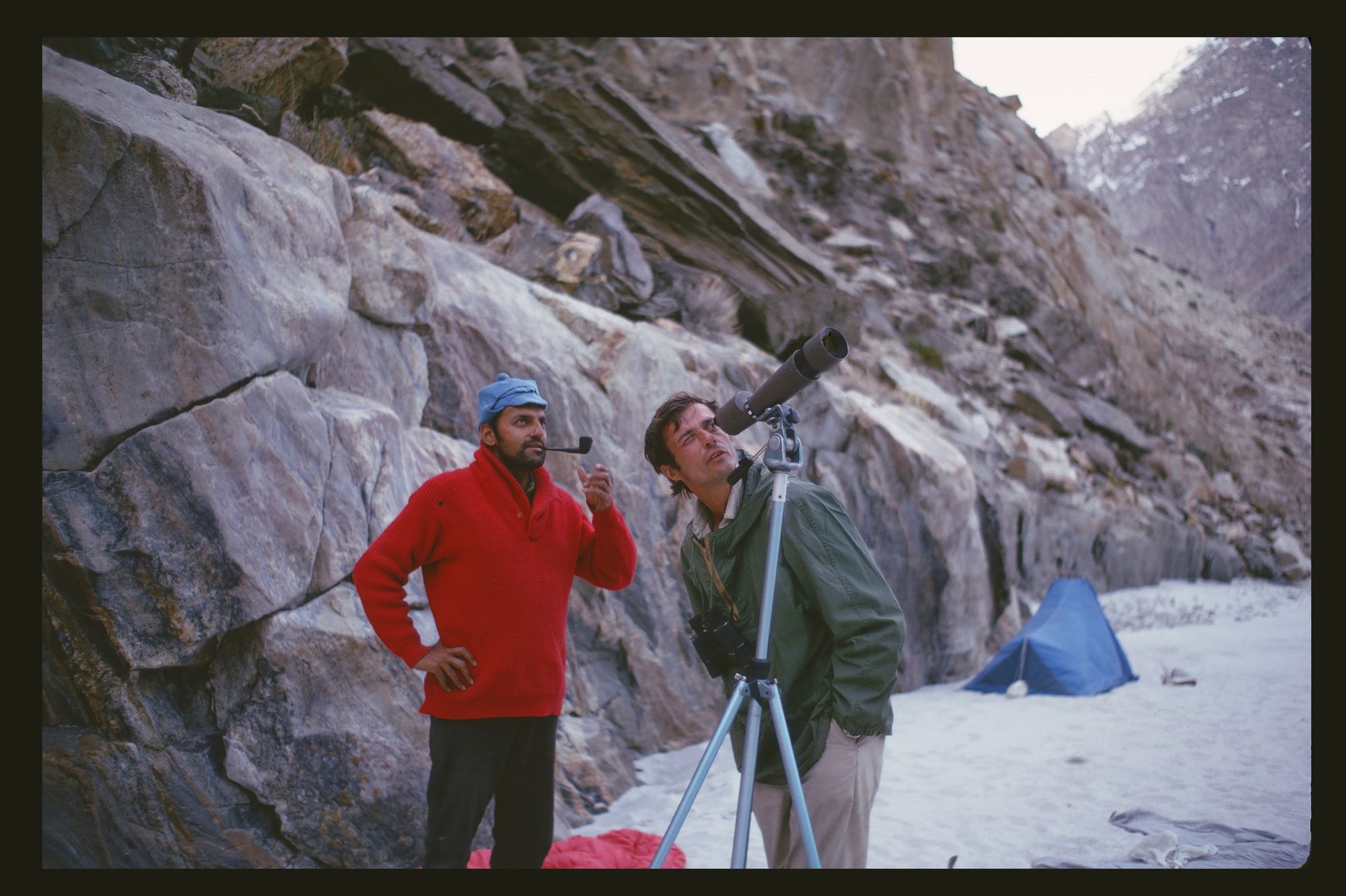 A man in a red sweater smokes a pipe and talks to George Schaller, who looks into a telescope.