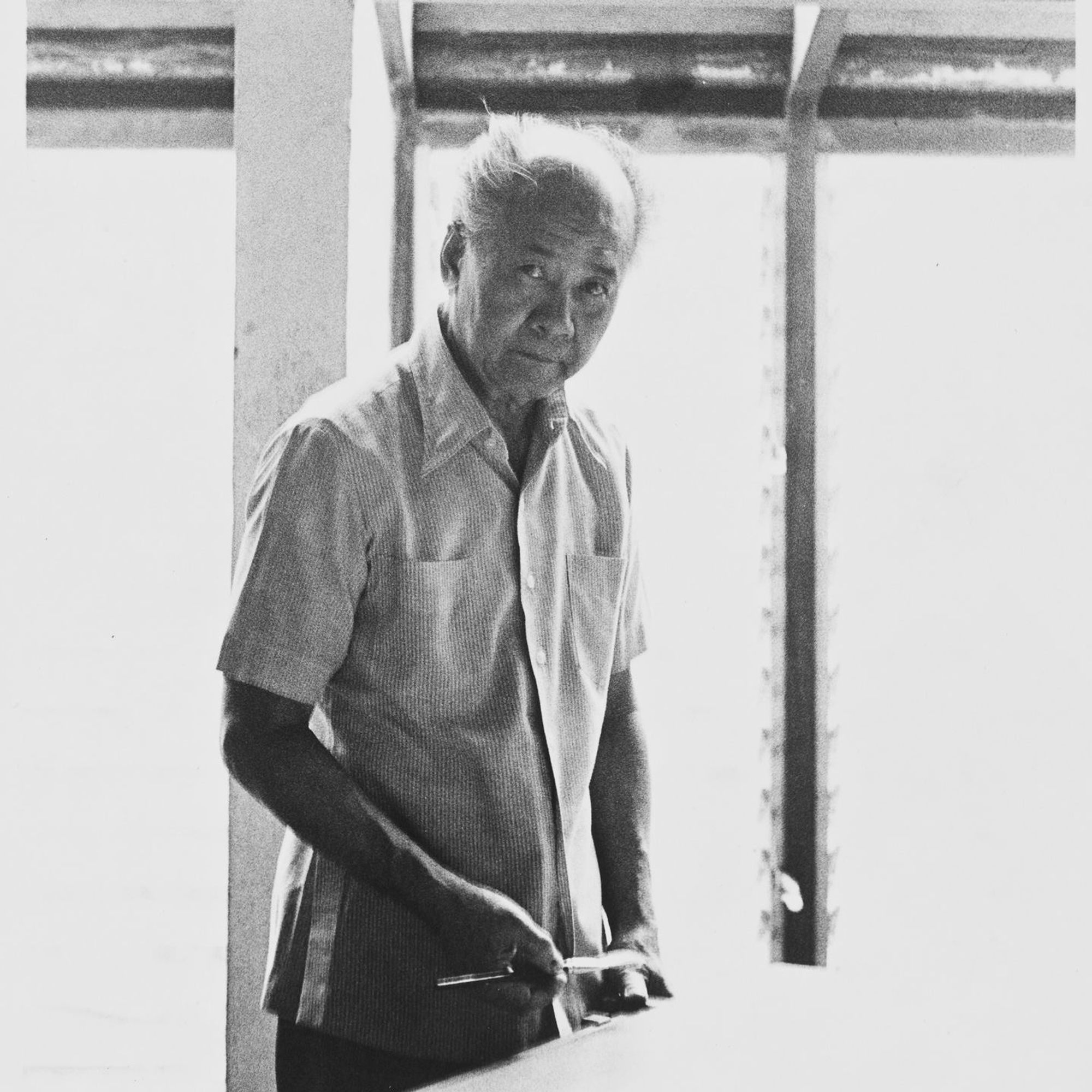 Bernardo stands at a table as an old man, gazing at the viewer.