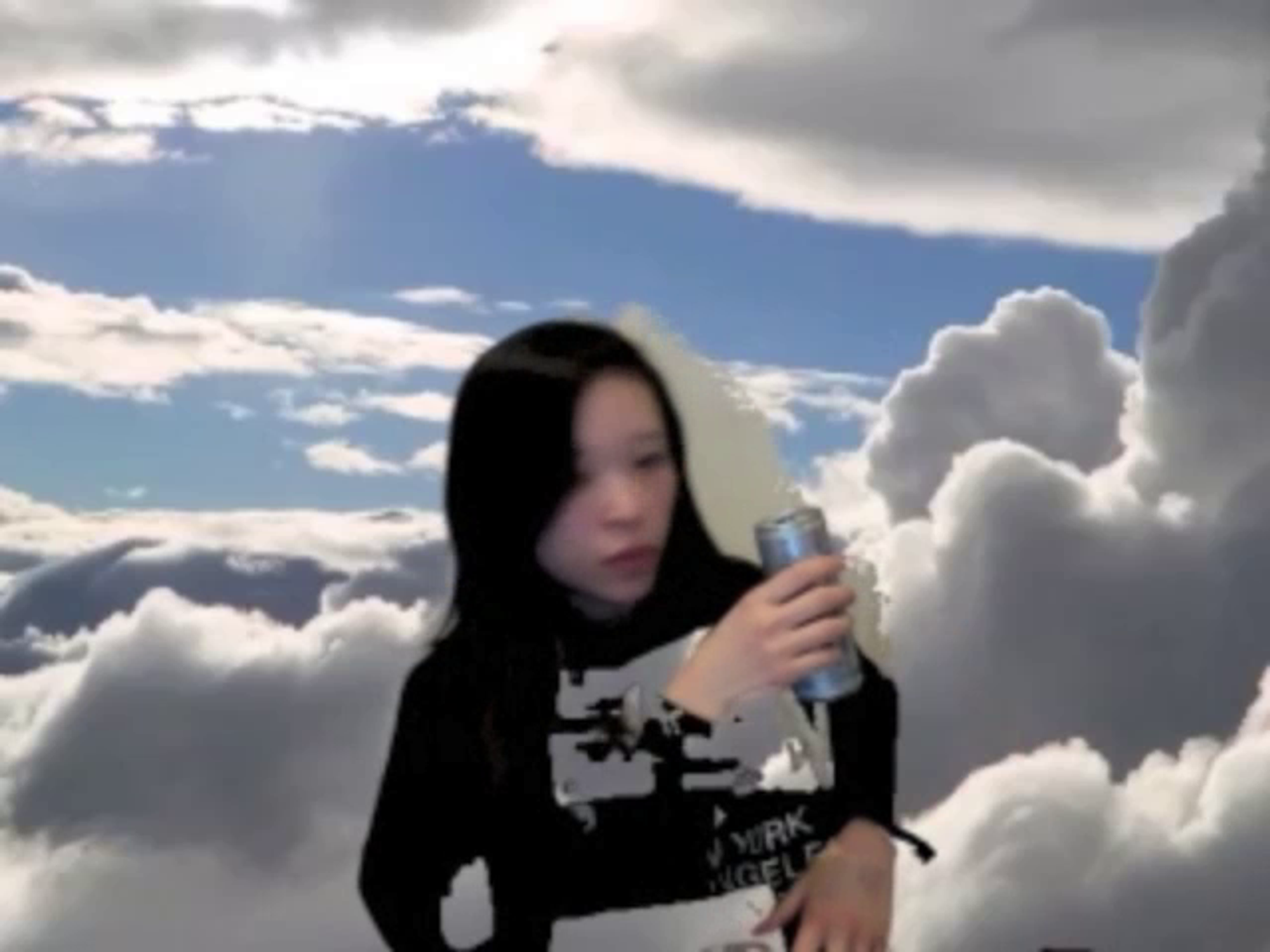 A webcam still with a woman wearing a black and white long sleeve shirt holding a Red Bull with a sky and clouds background