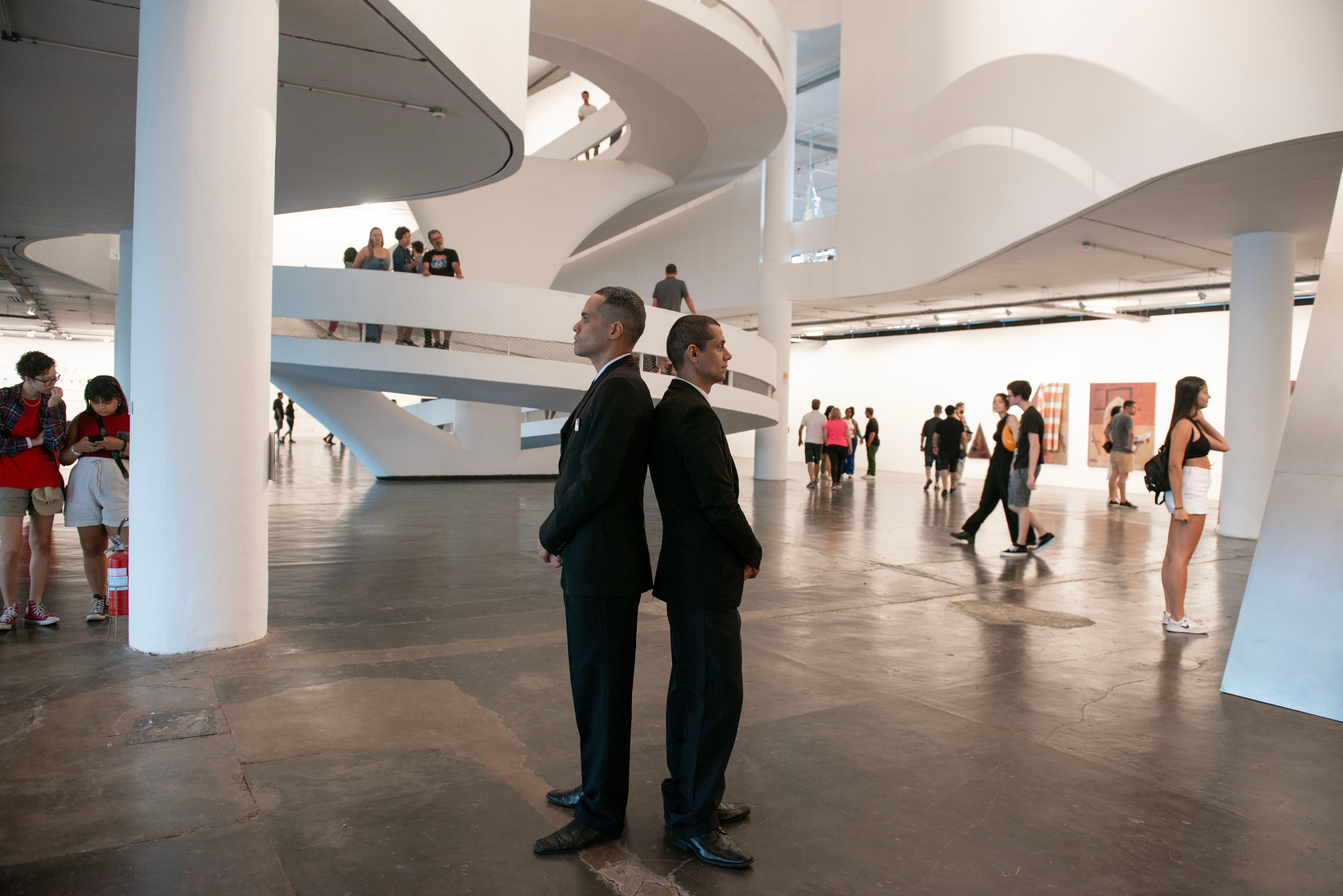 Two security guards stand back to back in a winding, white exhibition space packed with visitors.