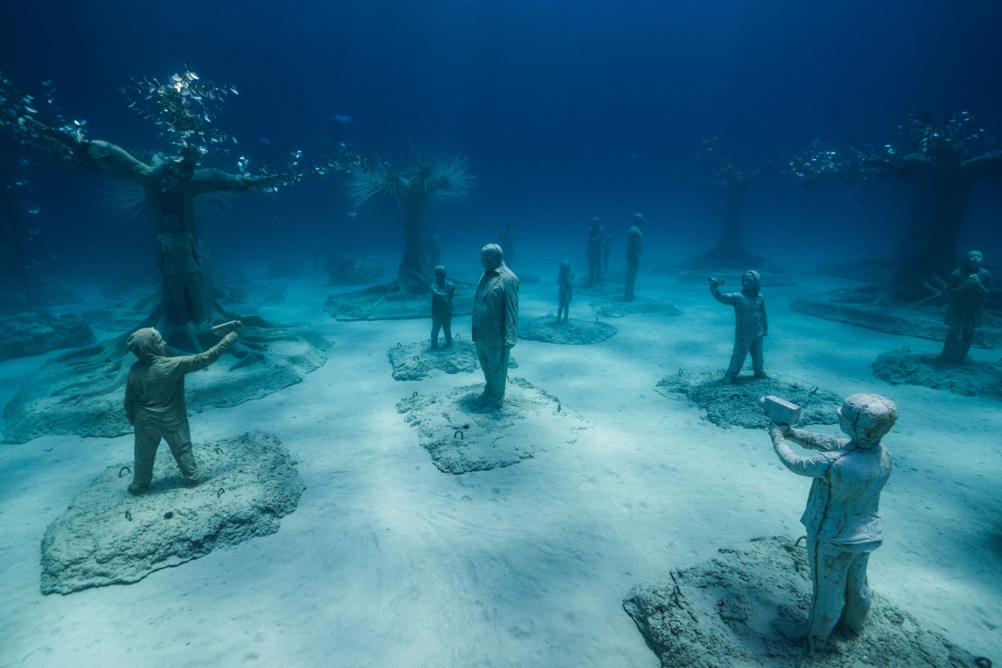 Human statues line the ocean floor, featuring children with cameras and a single adult figure at the center. 