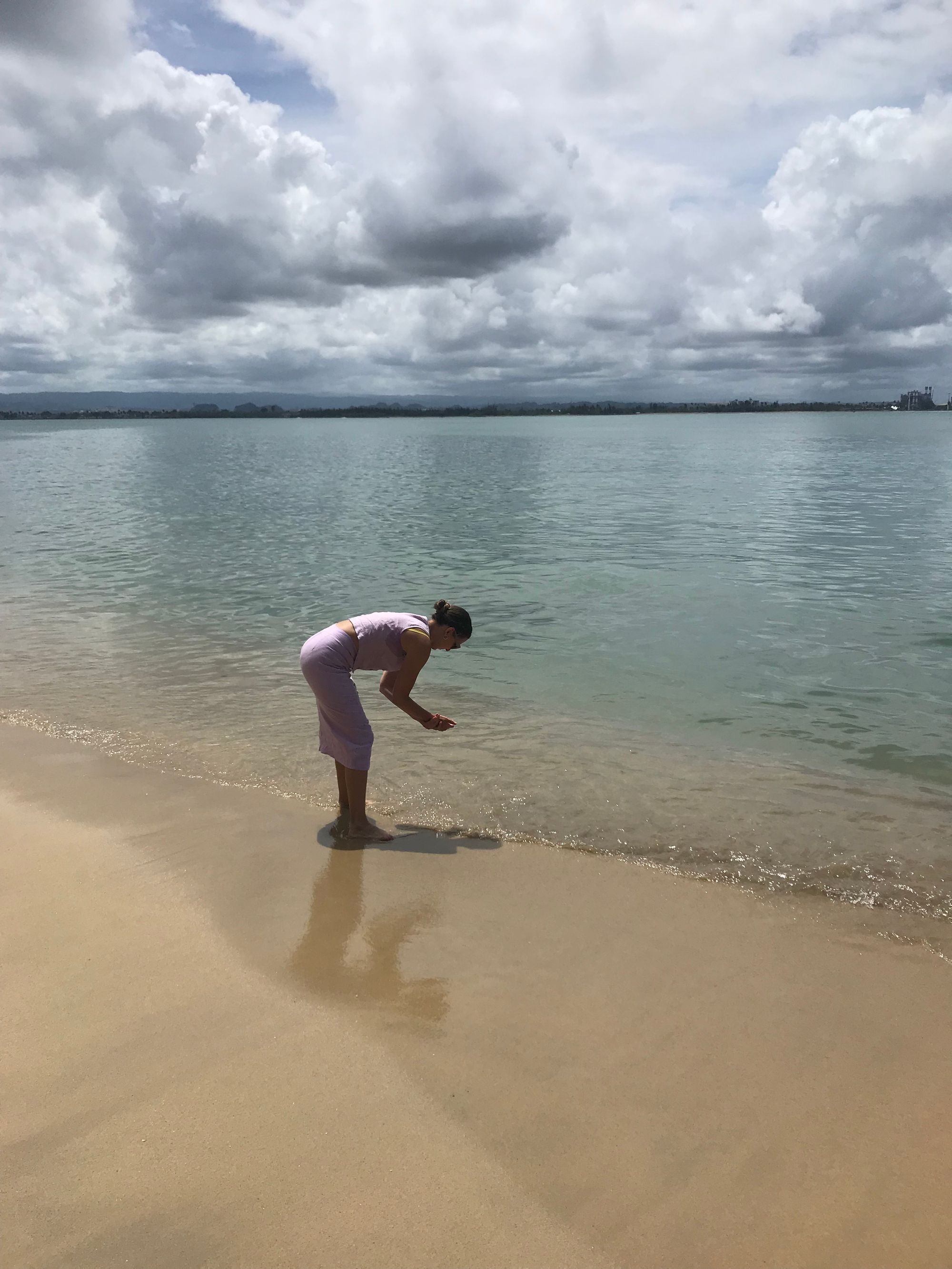 A woman wearing a skirt and tank top bends towards the seashore as she washes her hands in the ocean.