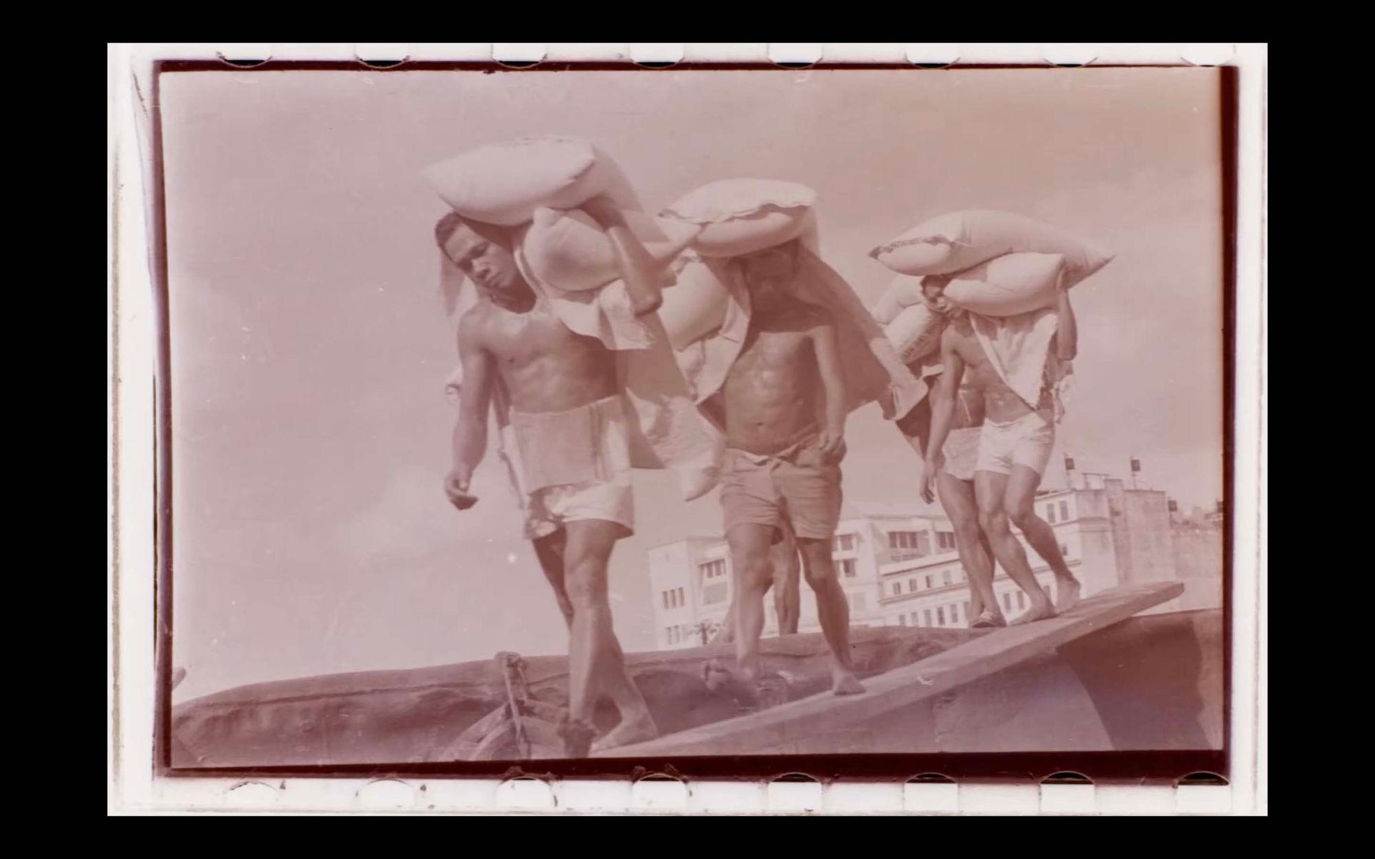 A sepia-toned photograph of three steverdores carrying sacks of rice. The sacks have soft, firm shapes and they rest on top and to the side of the stevedores' heads. The steverdores are shirtless and are wearing shorts.