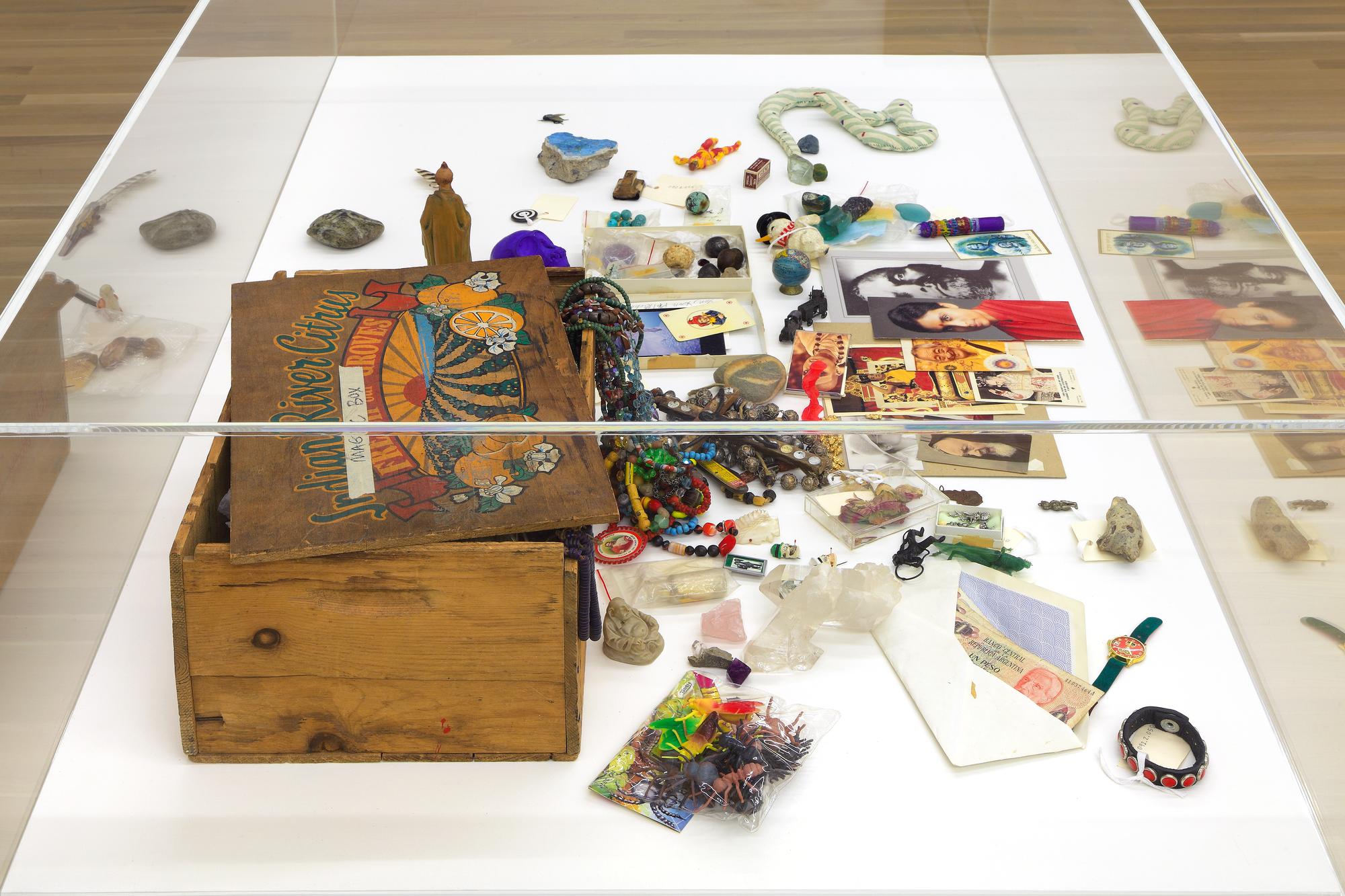A wooden box resting in a glass case, surrounded by colorful knickknacks: watches, photographs, jewelry, and the like.