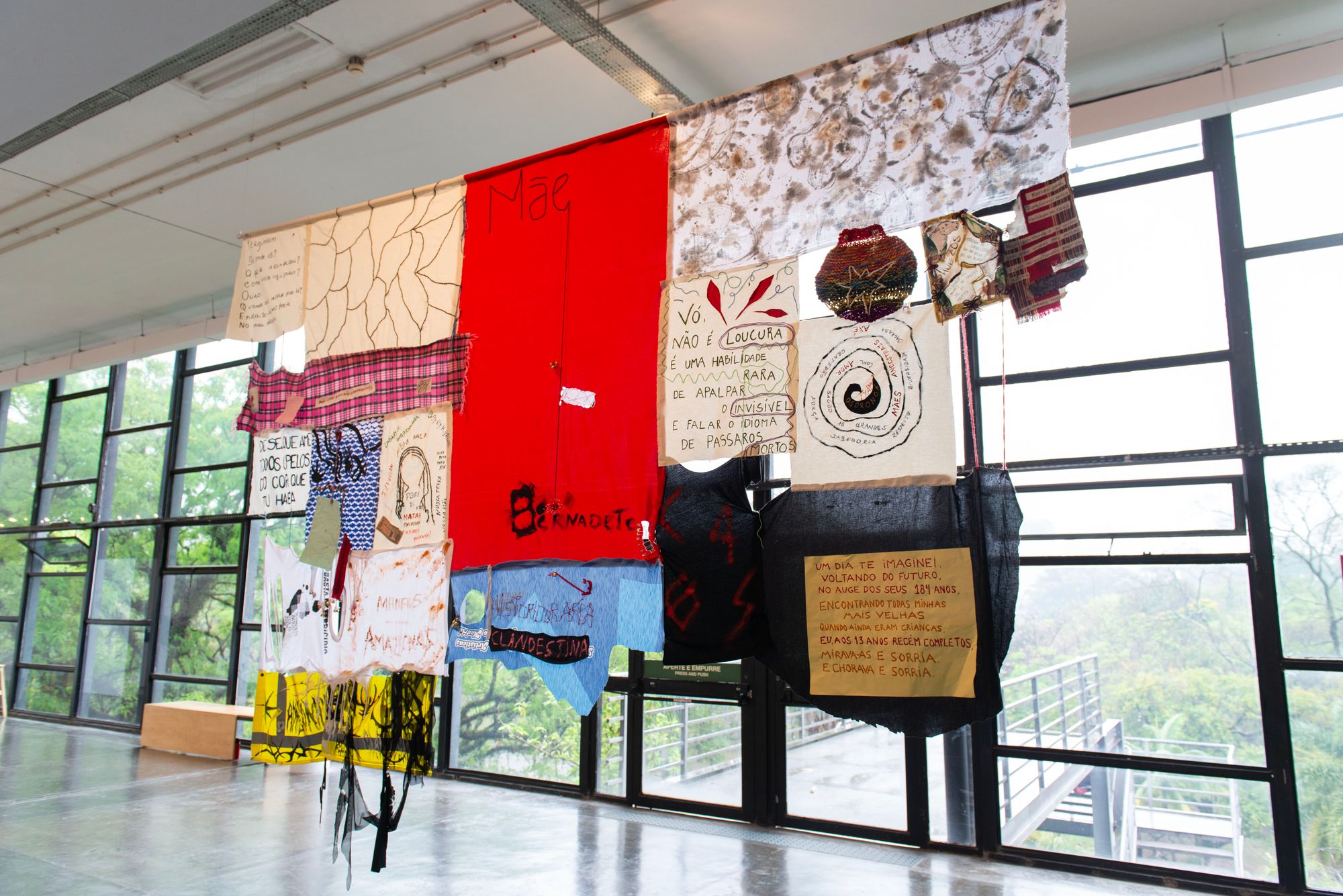 Quilted fabrics—with portugese text written across some fragments—hang from the ceiling in a windowed gallery space.