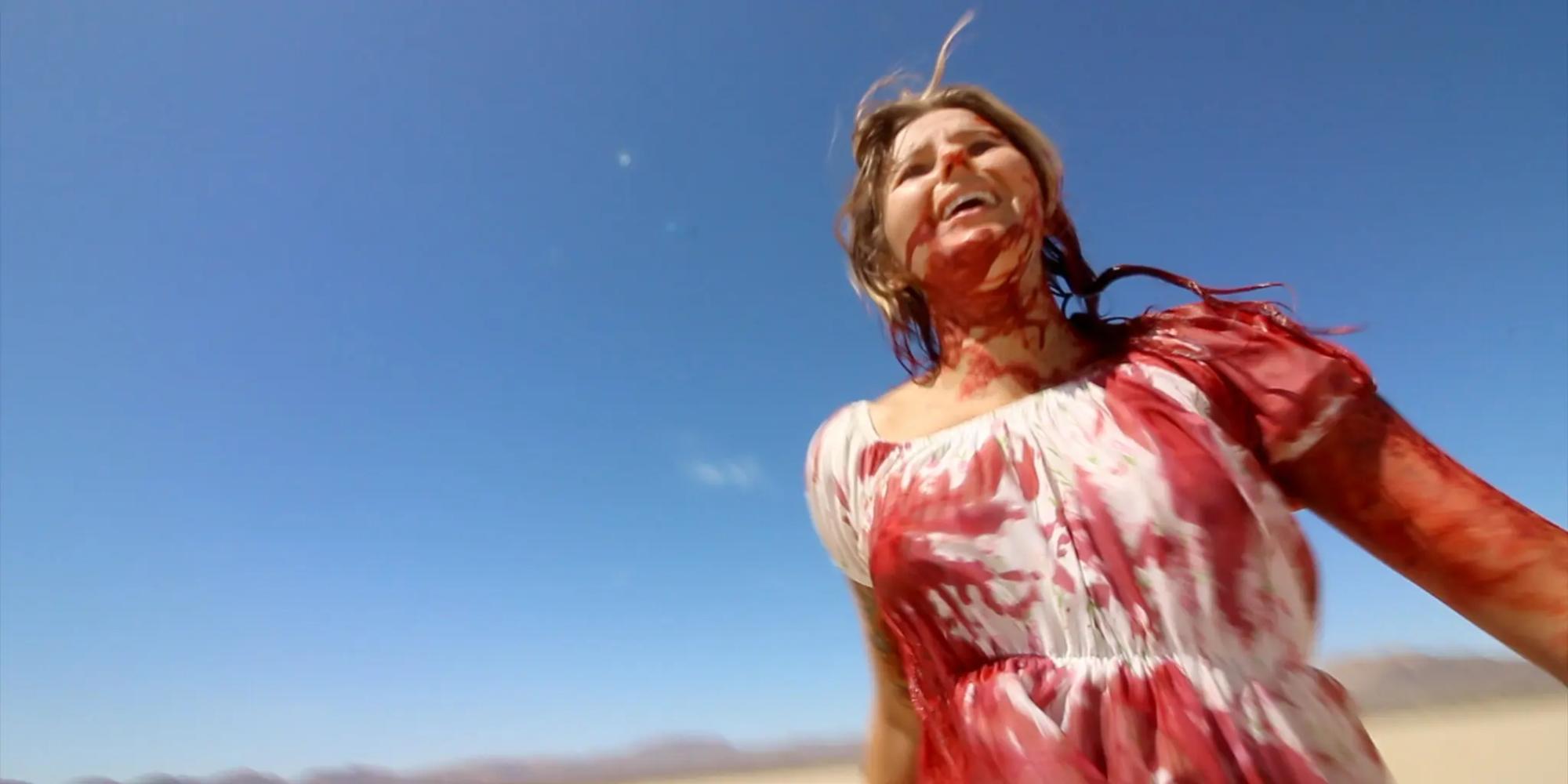 A blond woman runs through the desert in a white dress soaked with blood. 