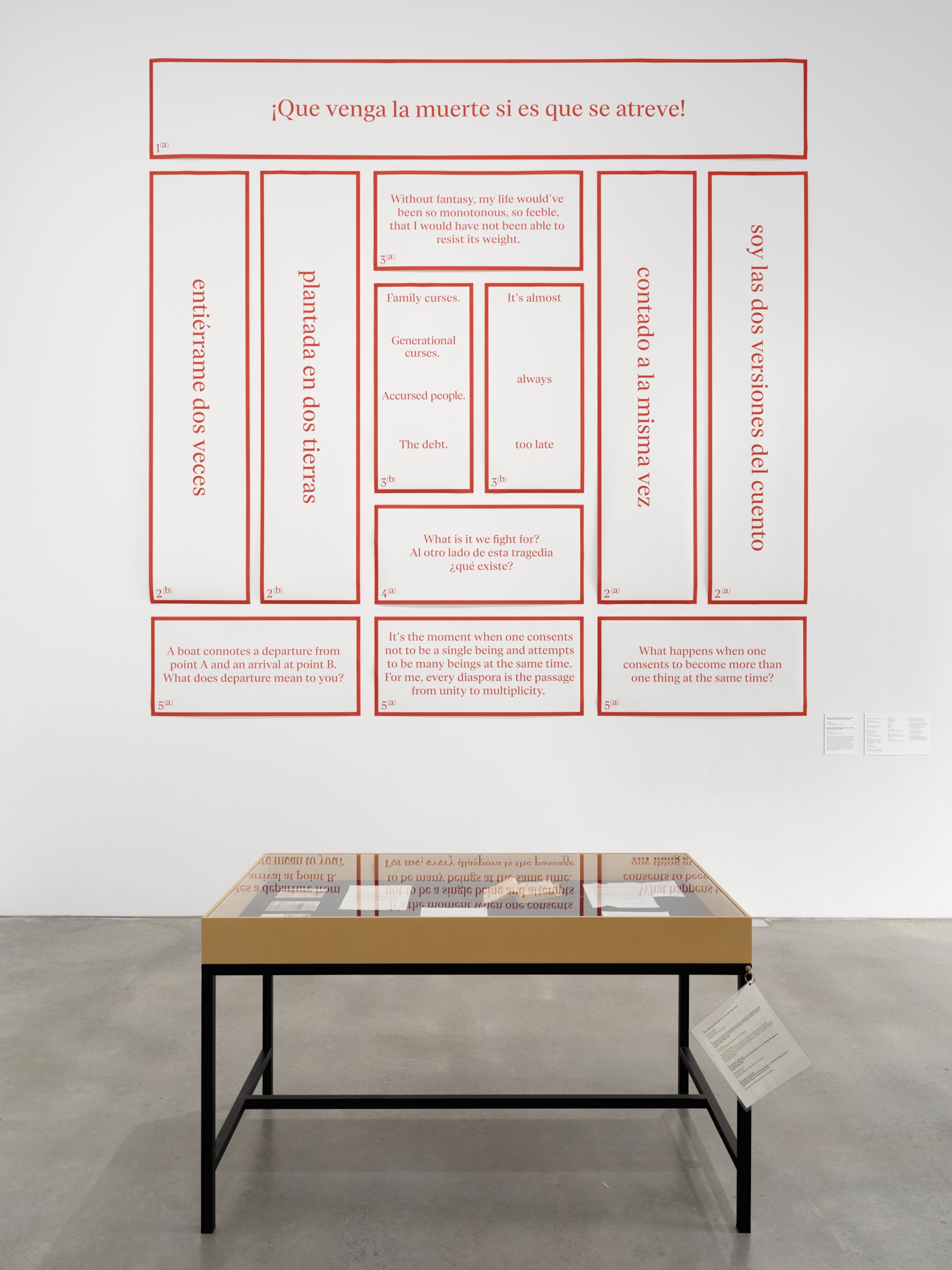 In a gallery space, an encased table showcases books and other ephemera, with an orange chart on the wall behind it.