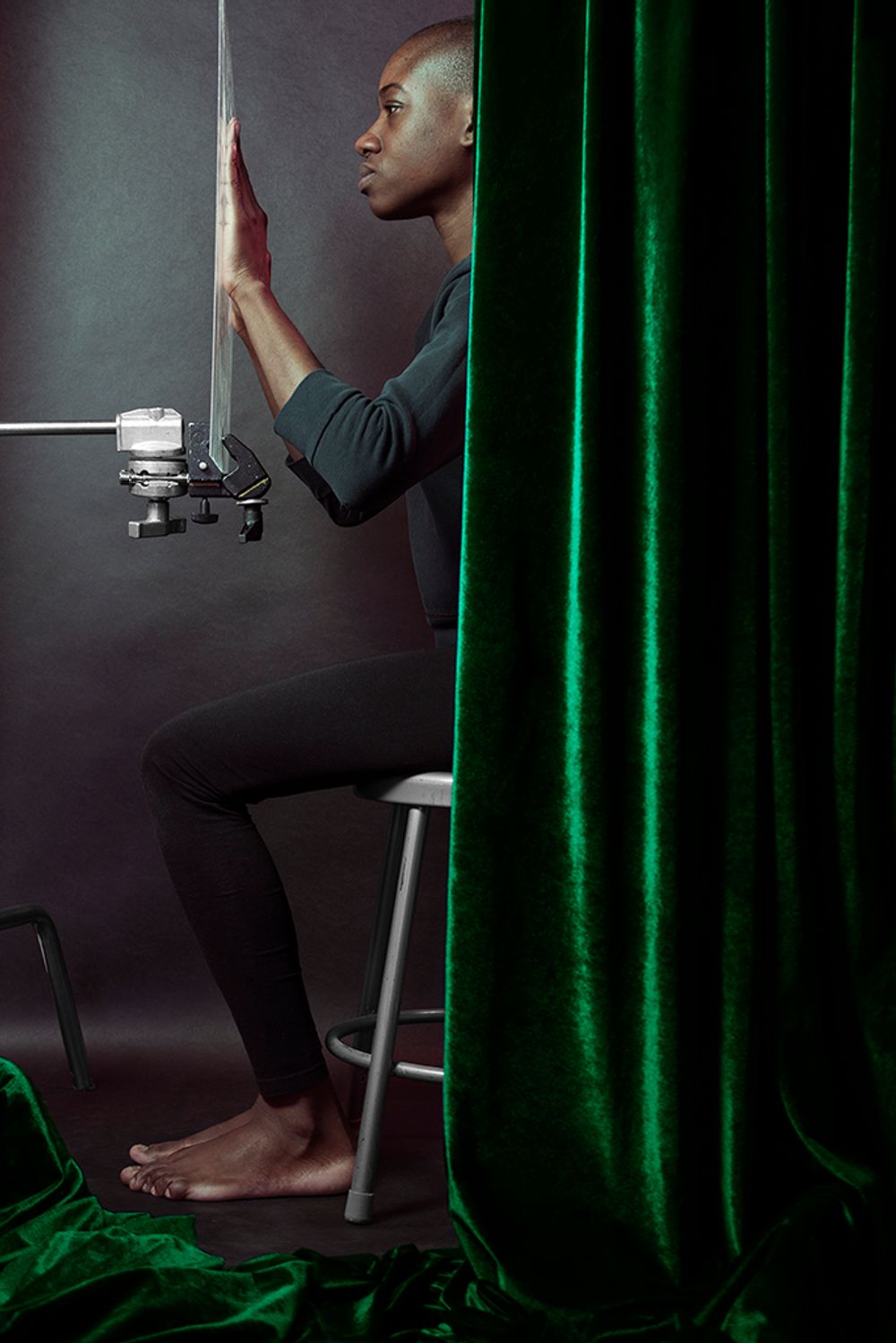 black person sitting on stool behind green curtain with hand flexed