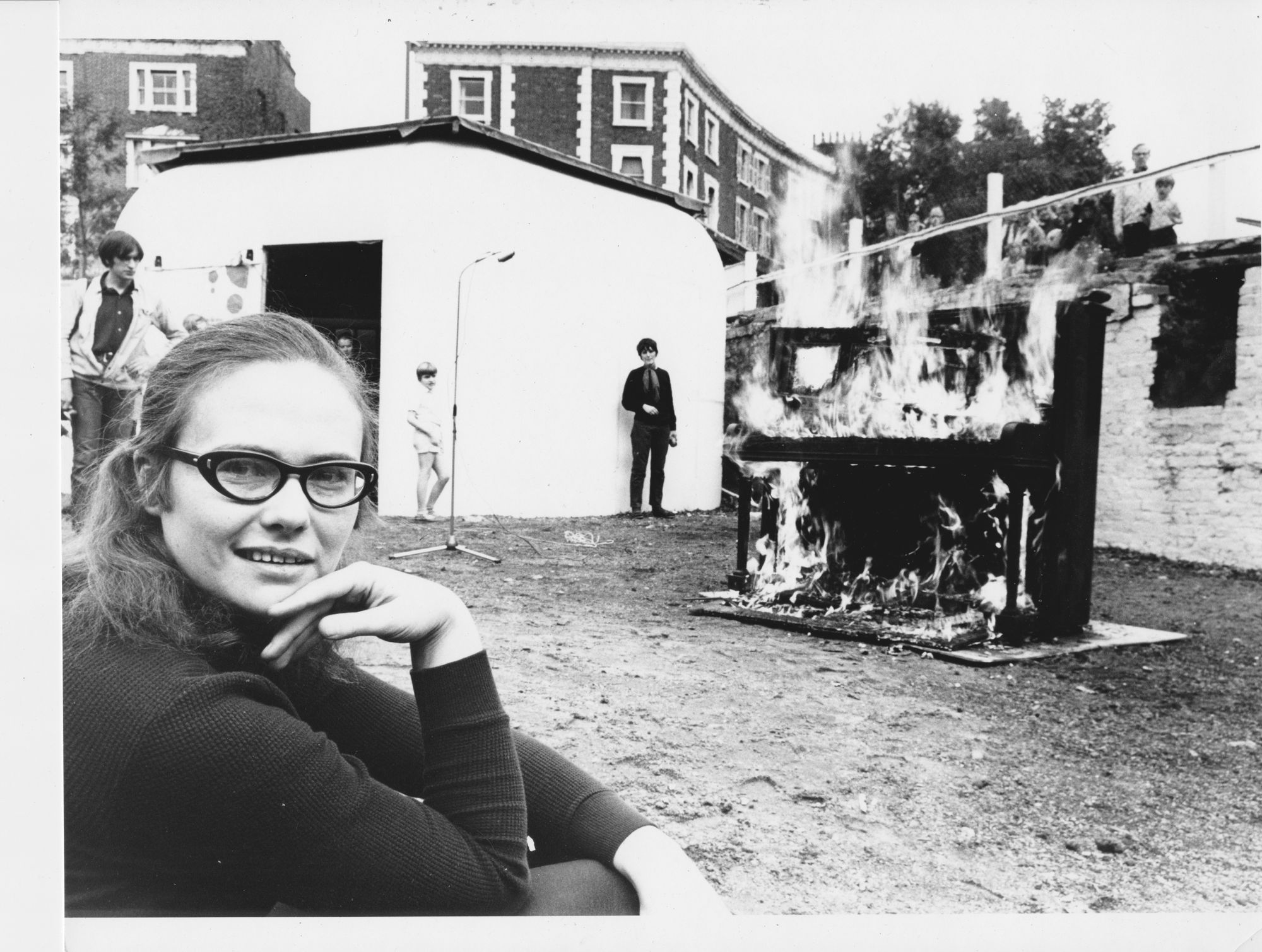 A black and white photograph of a young woman wearing glasses and looking away from a burning piano