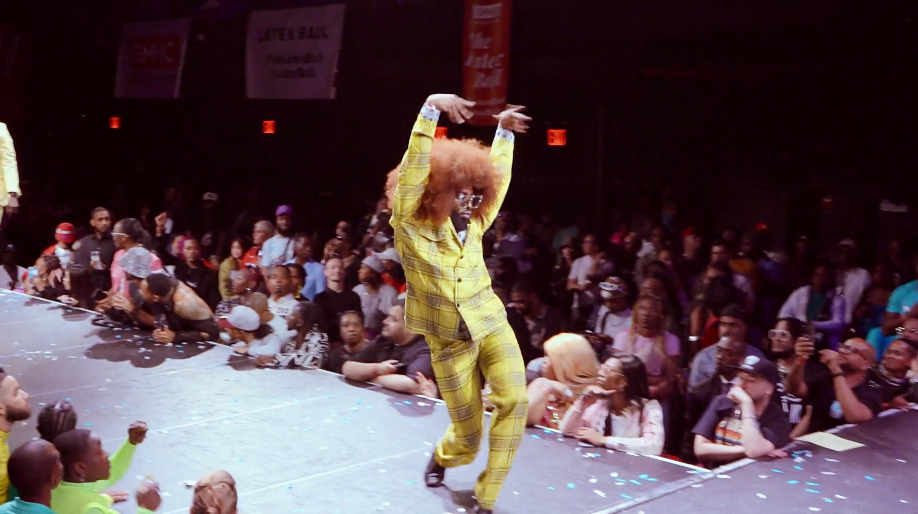 A drag queen strutting across a ballroom stage wearing a yellow plad suit