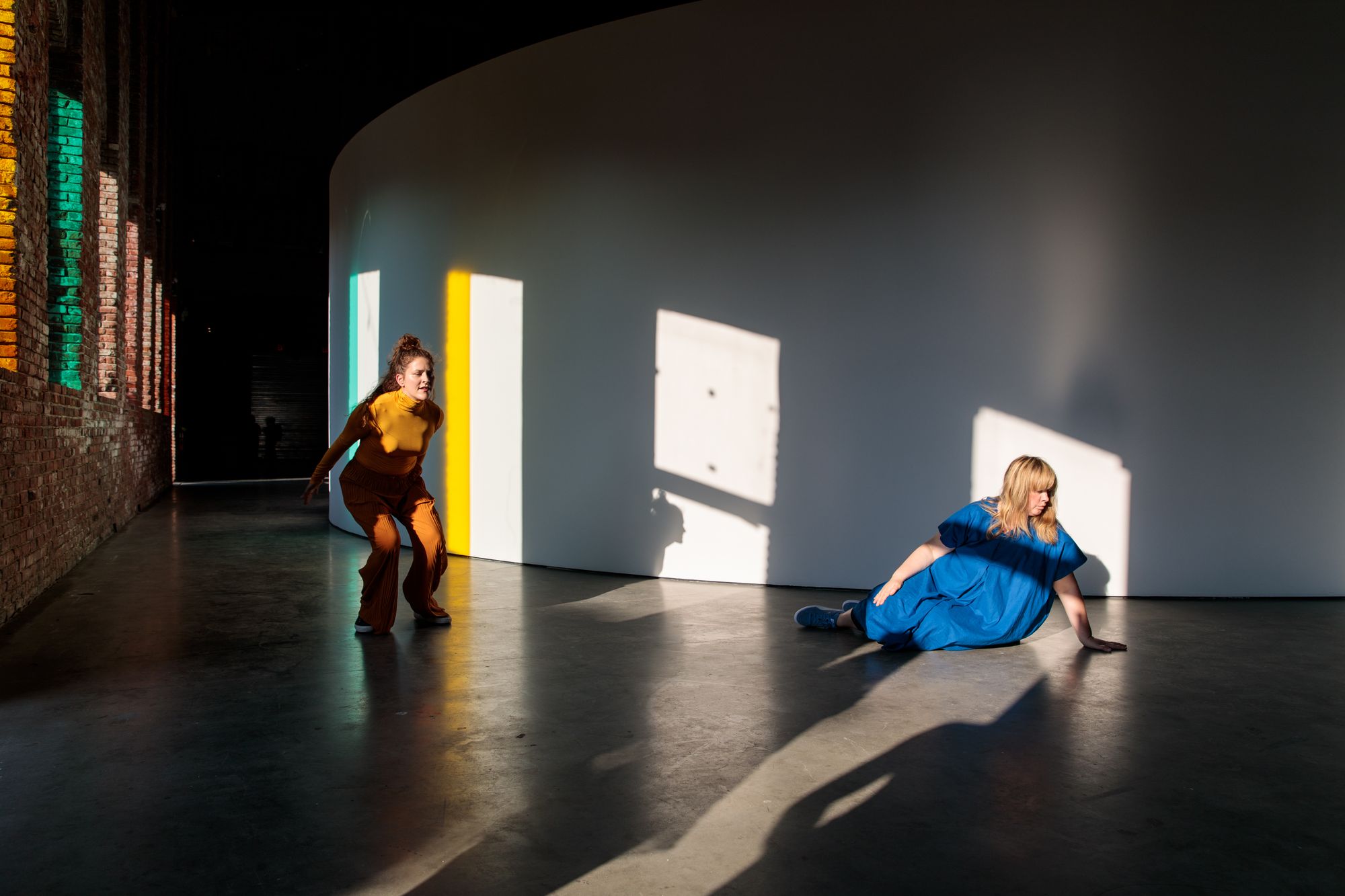 A woman in yellow crouches mid-movement, while a woman in blue taps her hand on her thigh, keeping beat.