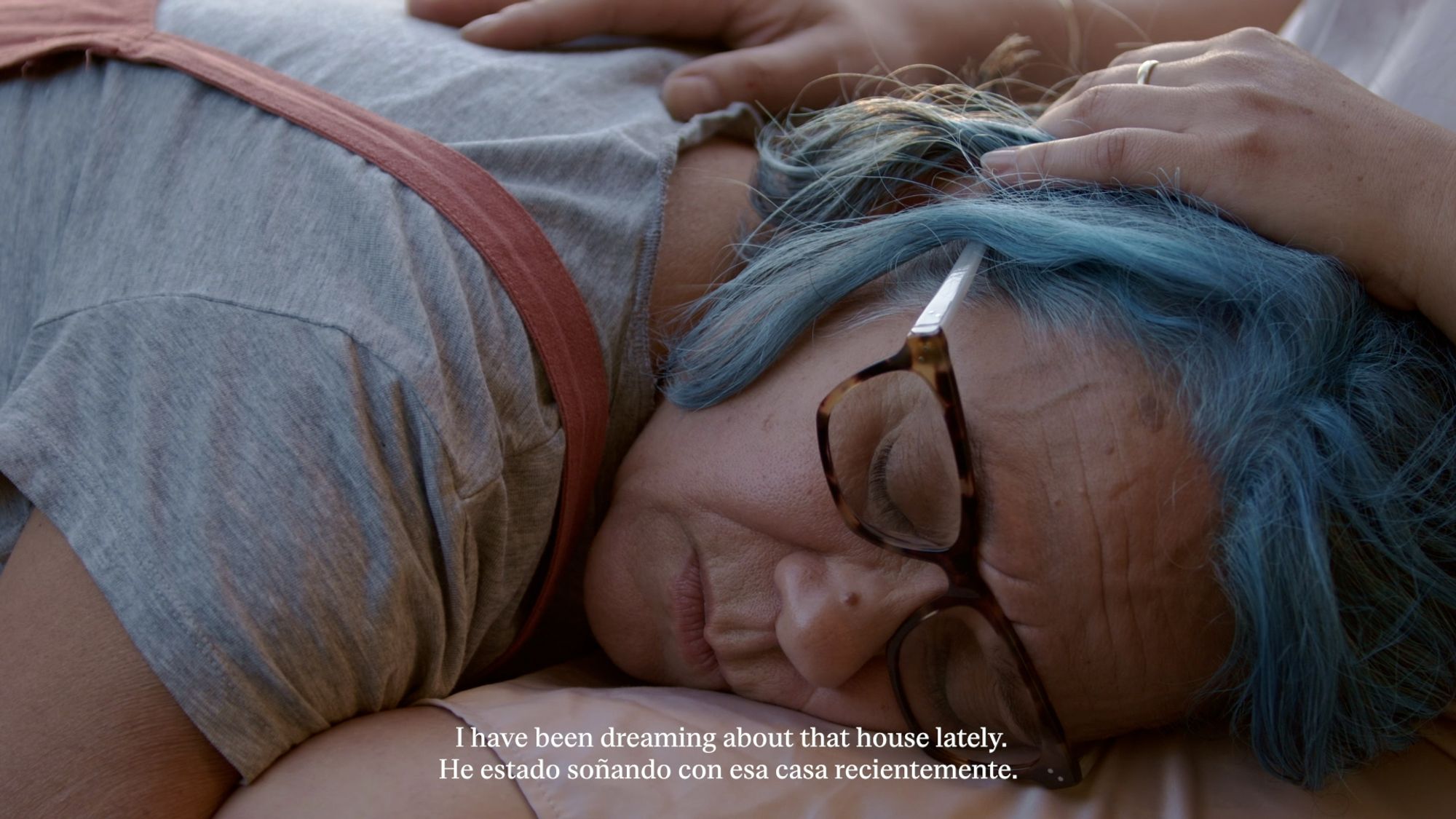 A film still of a woman lying with her head in someone's lap, with a subtitle at the bottom that reads: "I have been dreaming about that house lately" in english and spanish.