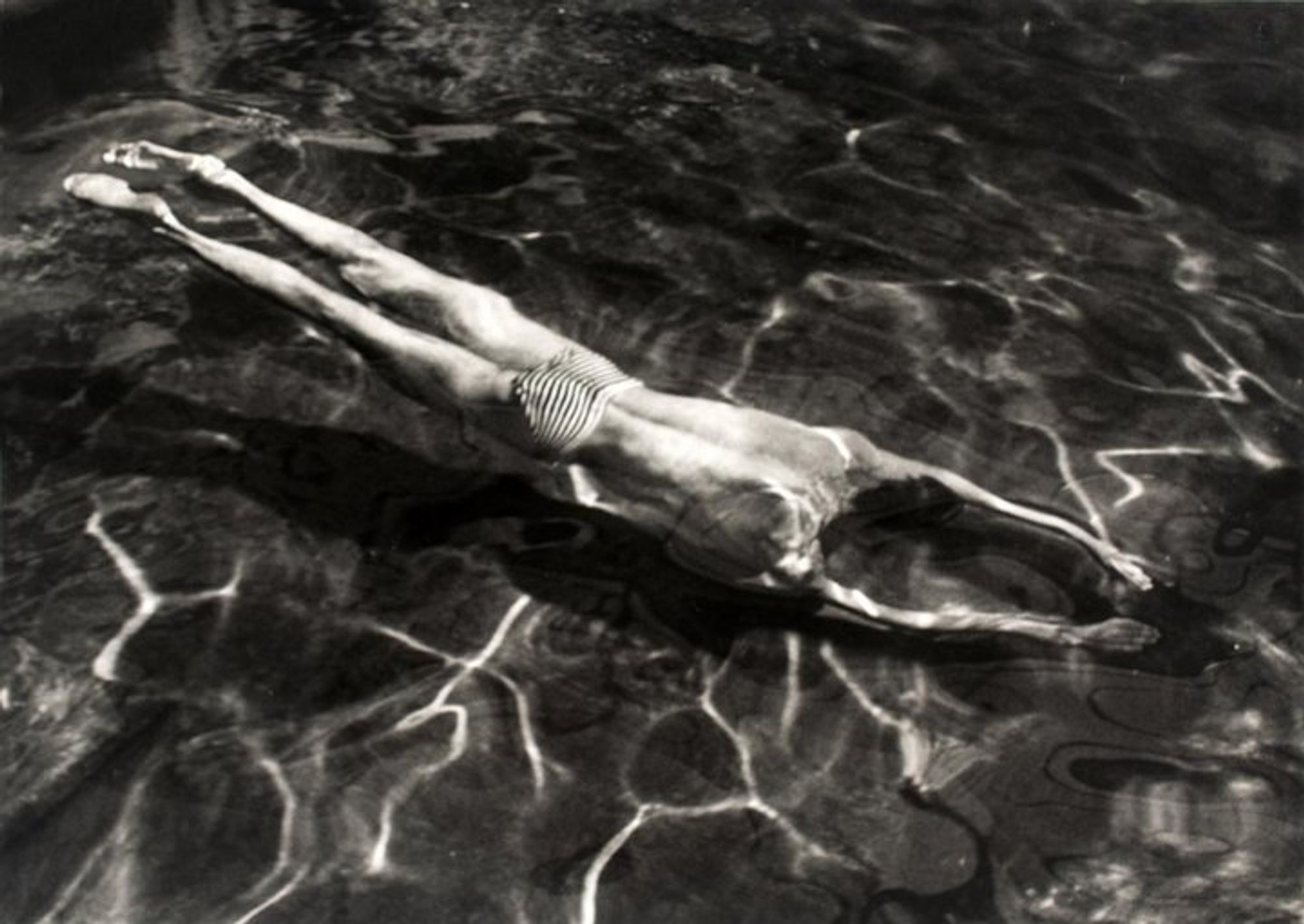 A black-and-white photograph of a swimmer underwater. The sun hitting the water forms squiggly lined patterns. The swimmer's arms are straight out in front of him and his body makes a long line.