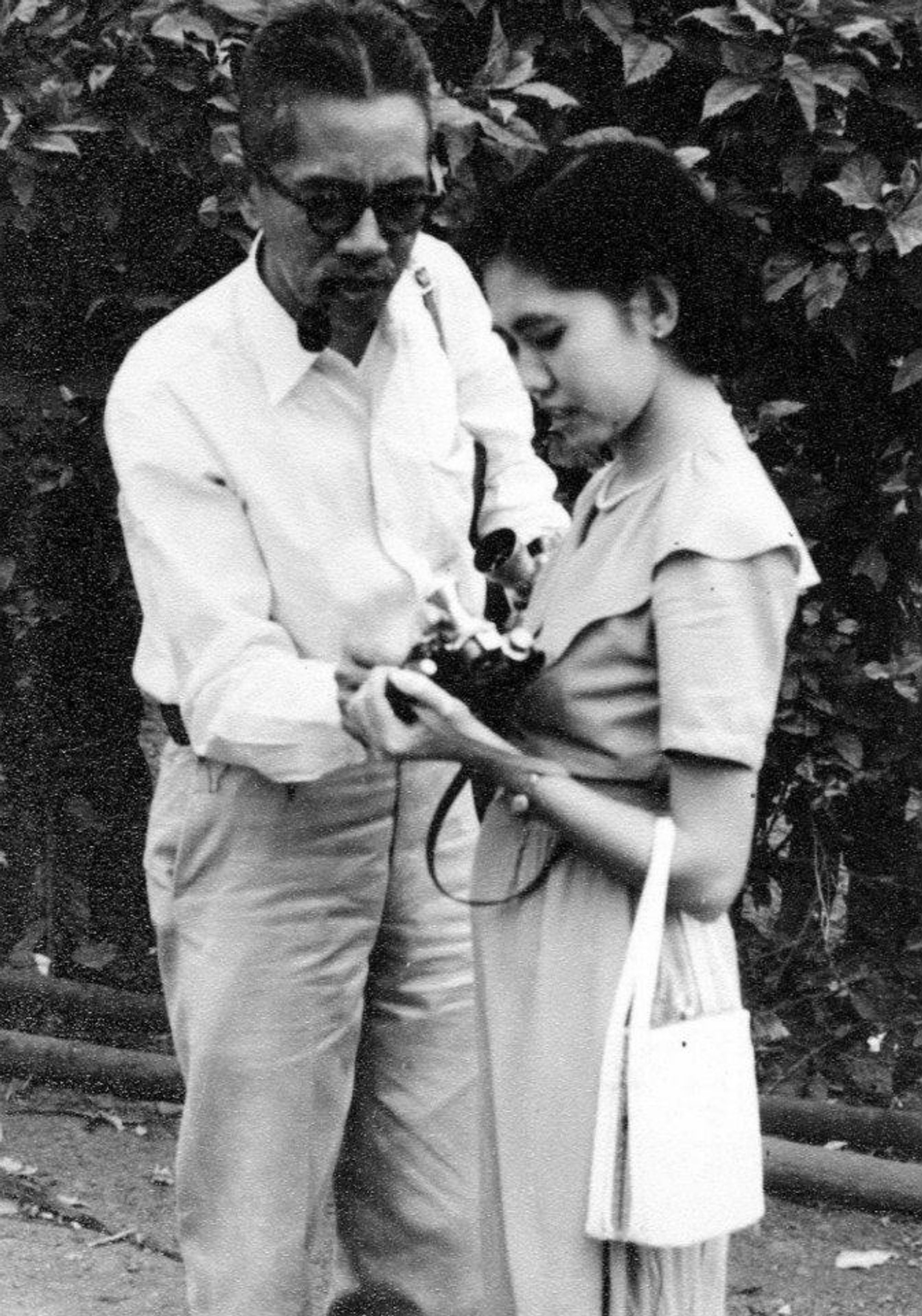 A black-and-white photograph of Protomartir and a woman sitting a camera that the woman is holding. He is wearing sunglasses and a white button-up shirt. She is wearing a short-sleeve dress and what looks to be a pearl necklace.
