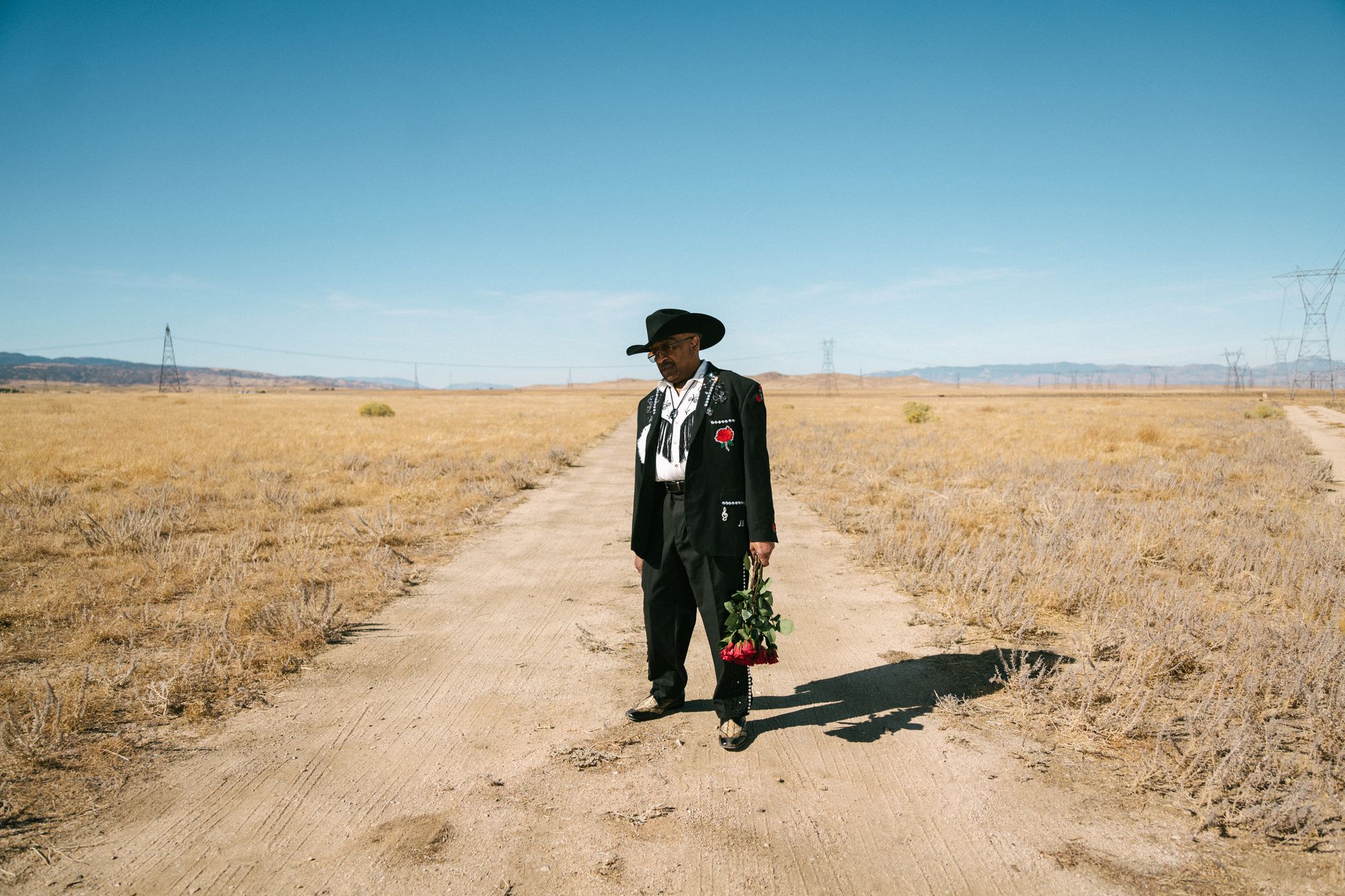 A black man, the musician Swamp Dogg, is shown standing on a sandy field. He is wearing a black charro outfit and a cowboy hat, and is holding roses