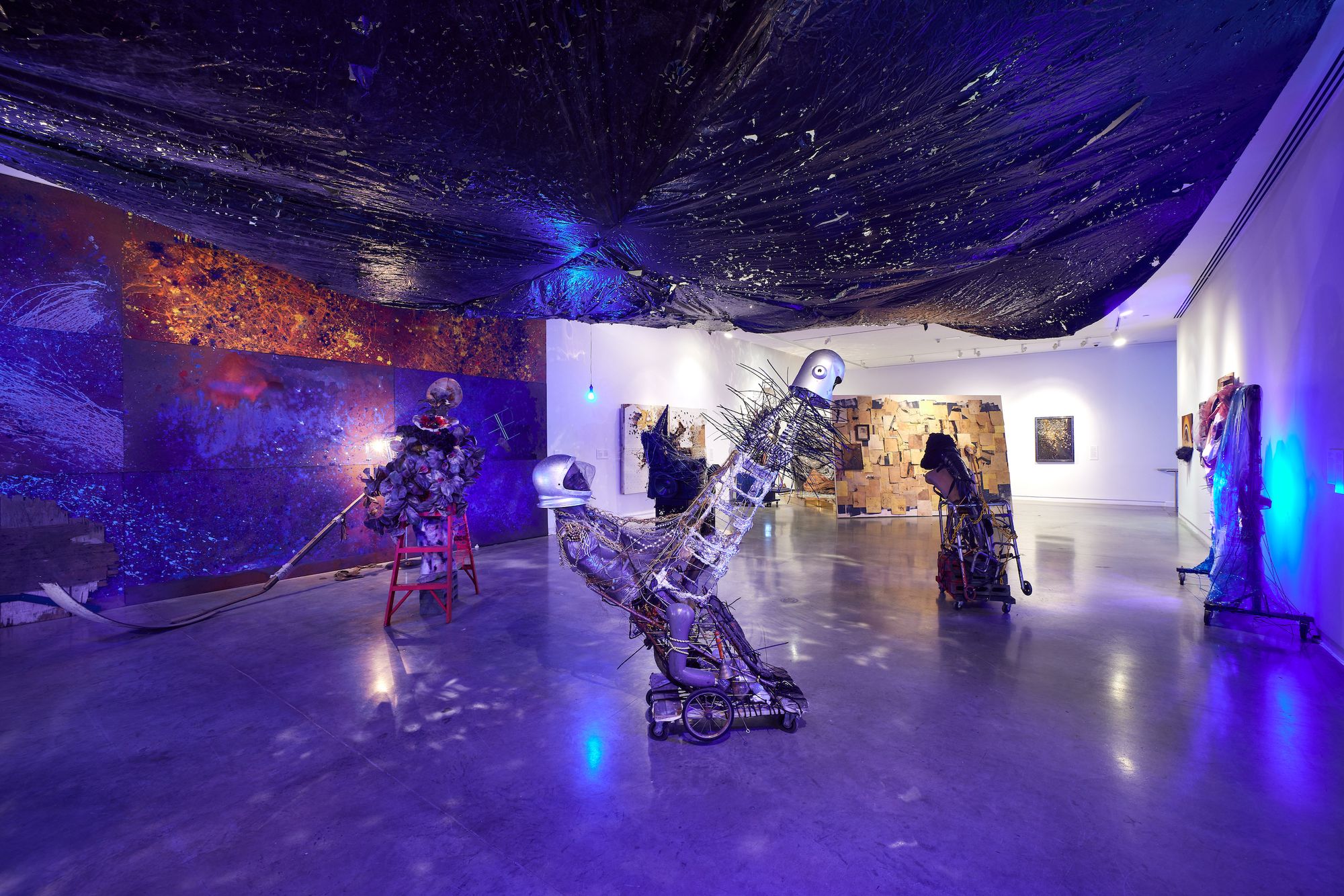 A gallery full of blue light, the ceiling draped in trash bags, with art pieces assembled from mixed metals and materials. 
