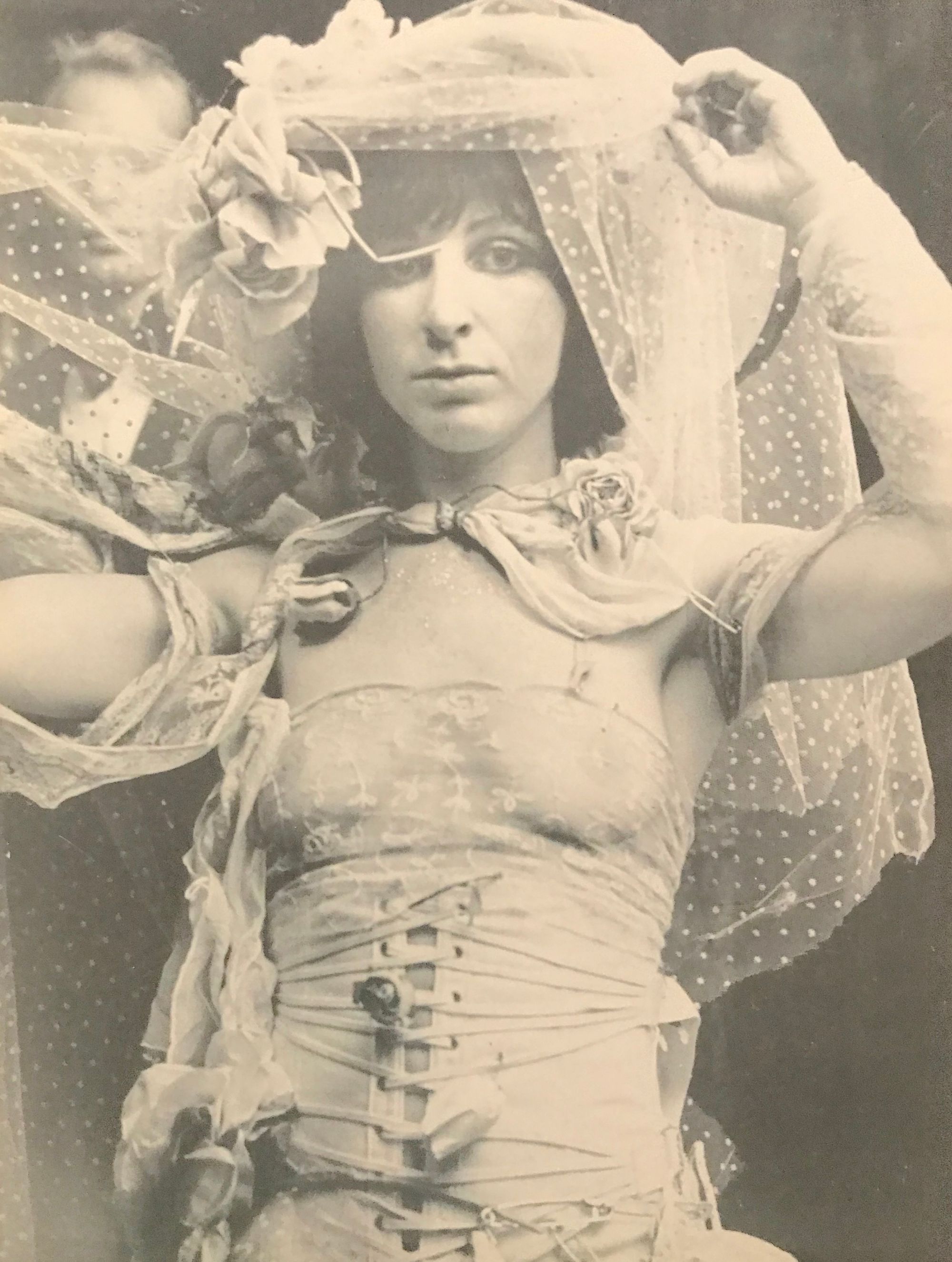 A faded black and white photograph of a woman covered in white lace, wearing a corset top.