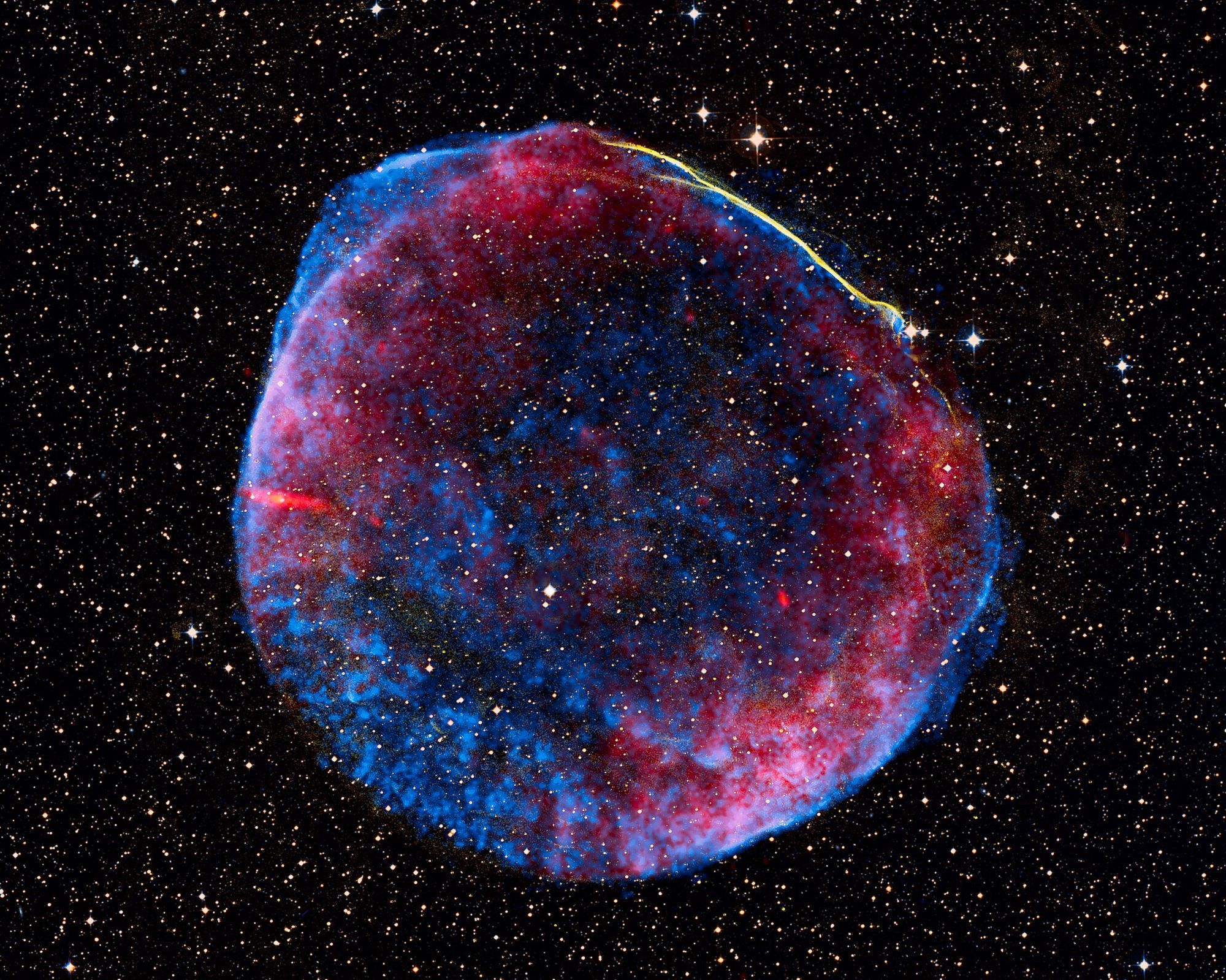 The hot remains of a supernova, resembling a maroon and blue bubble floating amongst thousands of stars in space.